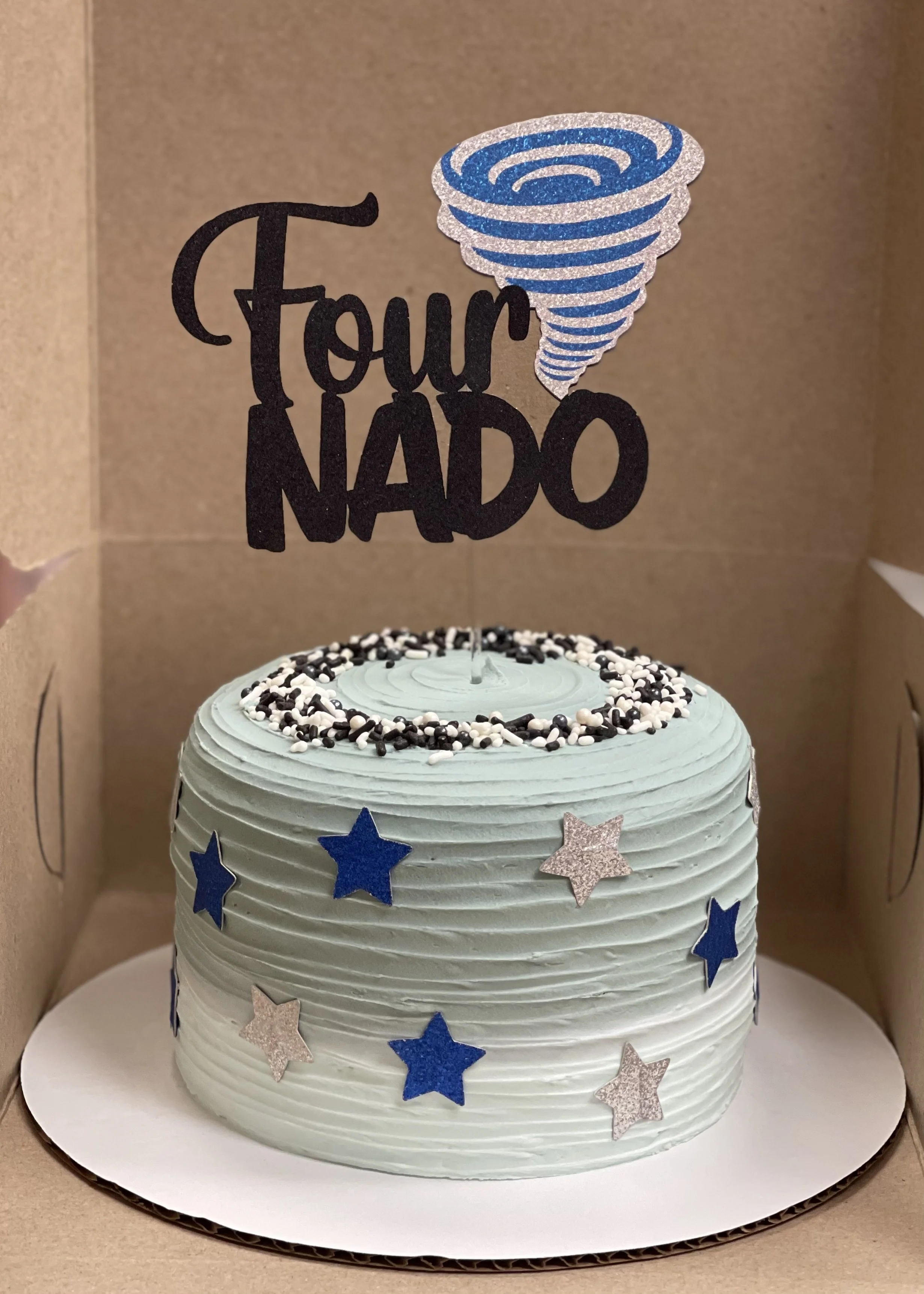 A light blue birthday cake decorated with silver and blue star-shaped sprinkles inside a cardboard box with a blue tornado cake topper and the words 'Four Nado' in black text.
