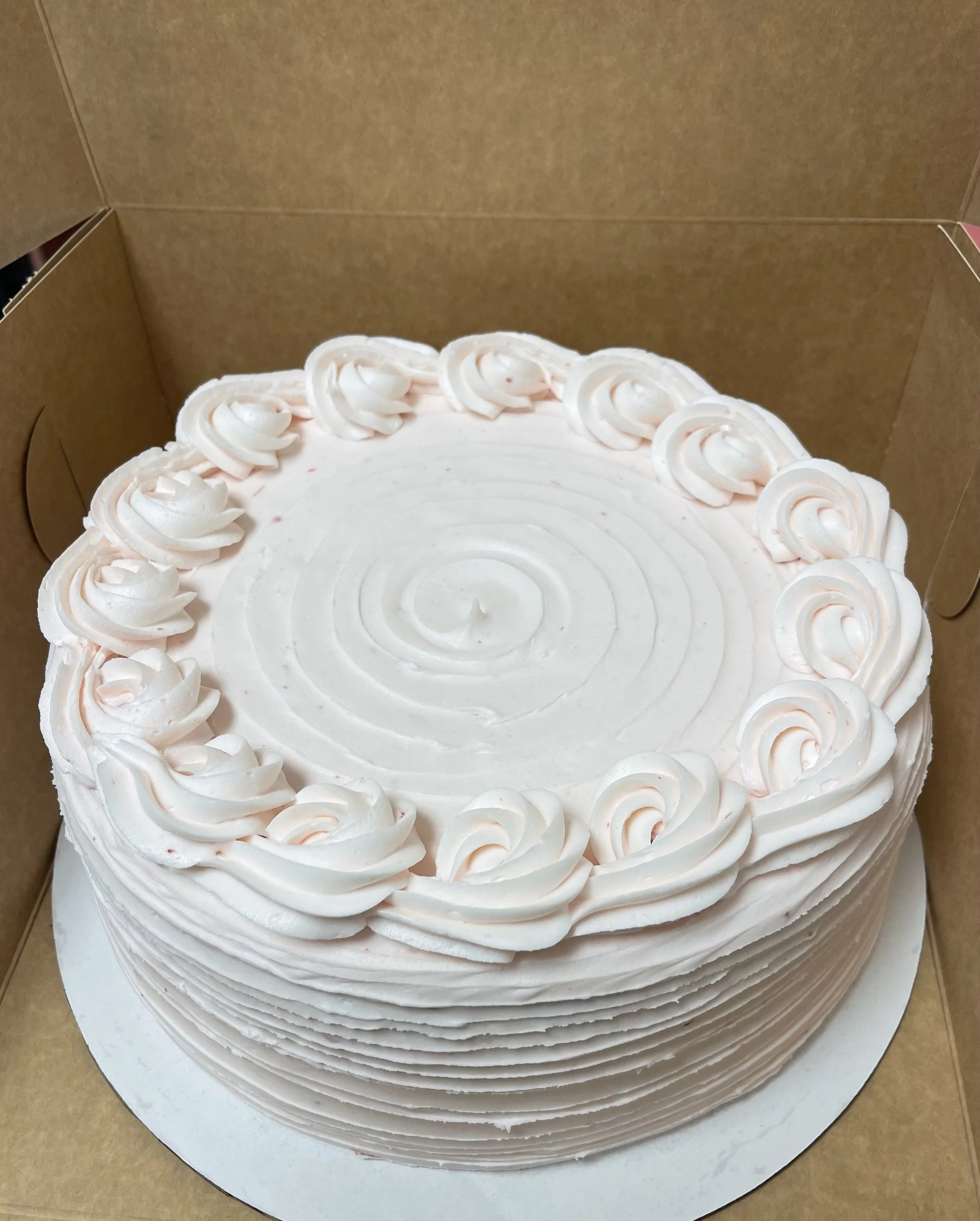A round, white frosted cake with pink piping on top, inside a cardboard bakery box.