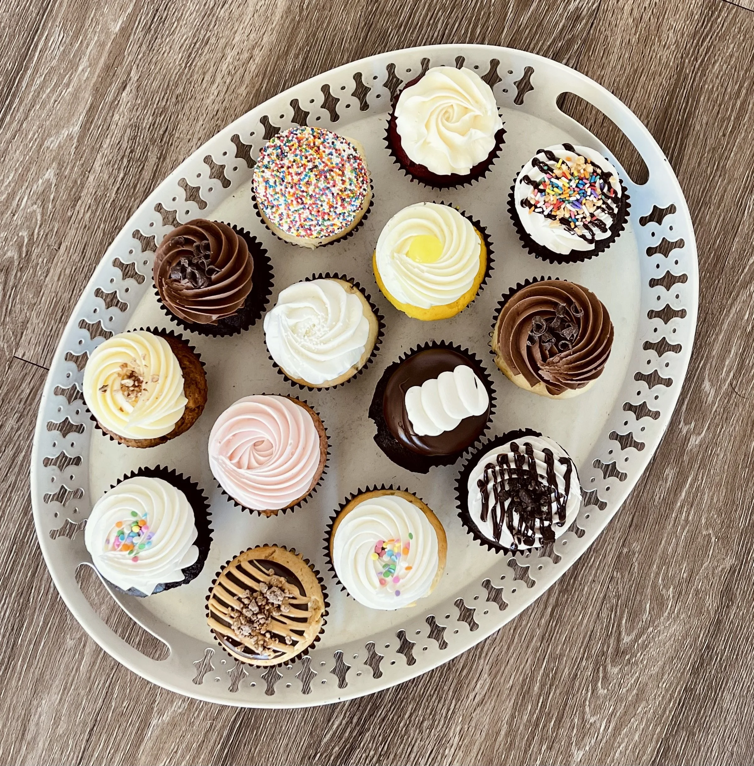 Tray of assorted decorated cupcakes with various frosting and sprinkles on a wooden surface.