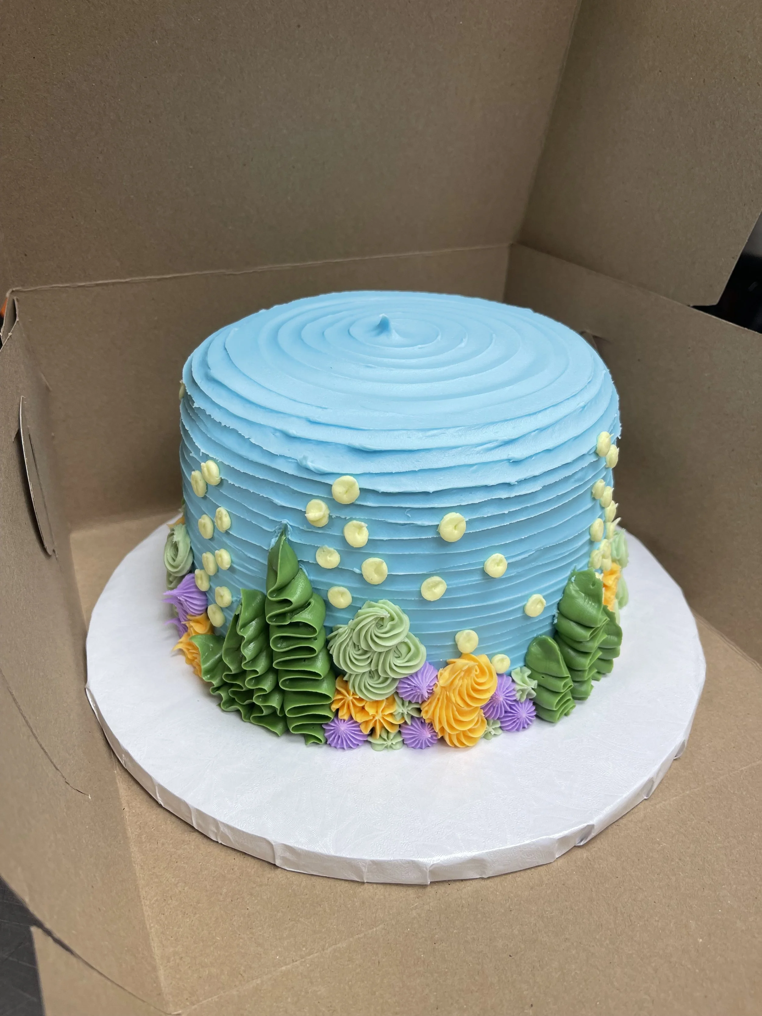 A colorful round cake with blue frosting and piped floral decorations in green, yellow, purple, and orange, placed inside a cardboard box.