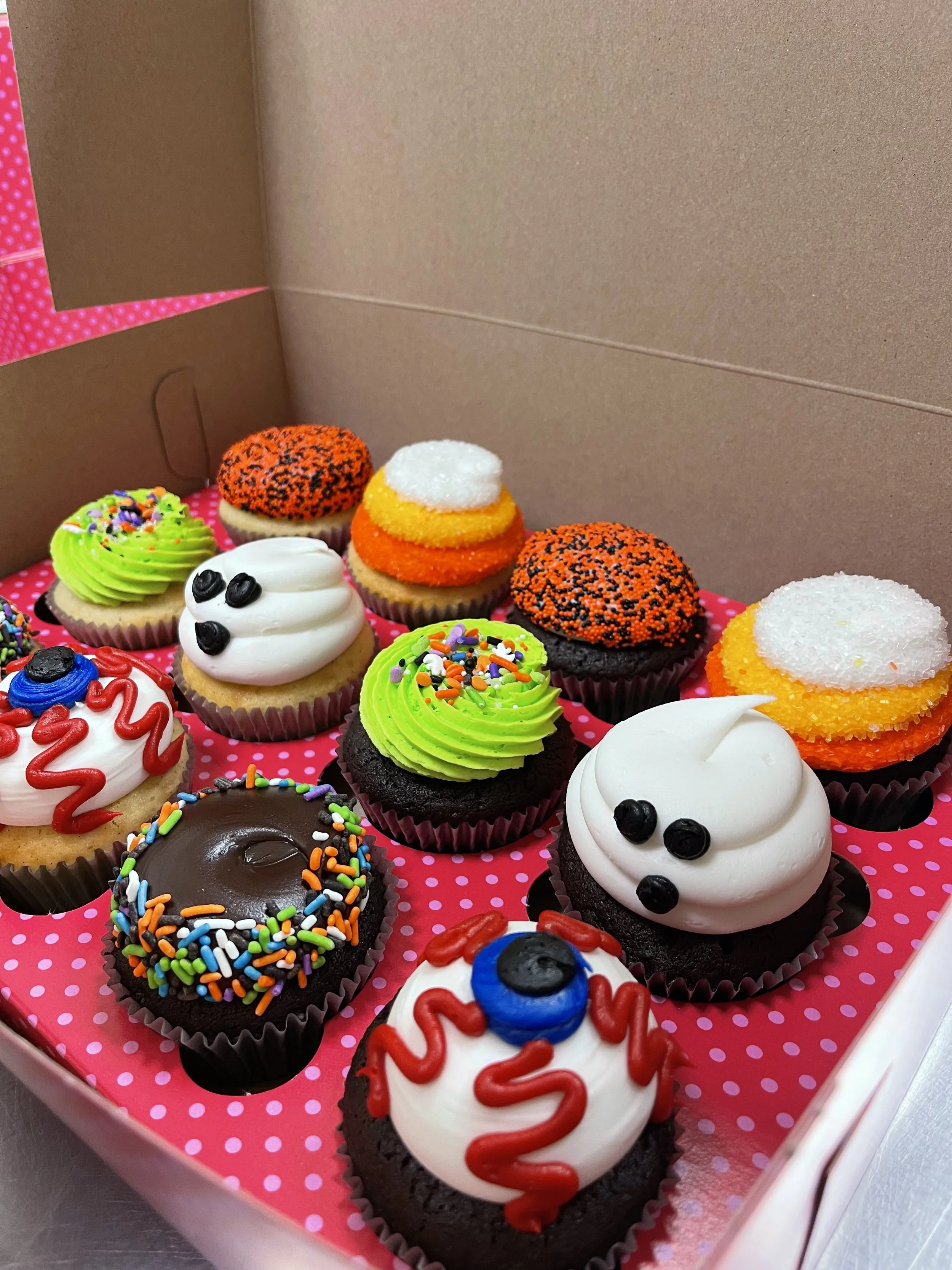 Assorted Halloween-themed cupcakes with colorful frosting, sprinkles, and decorations in a pink polka dot box.