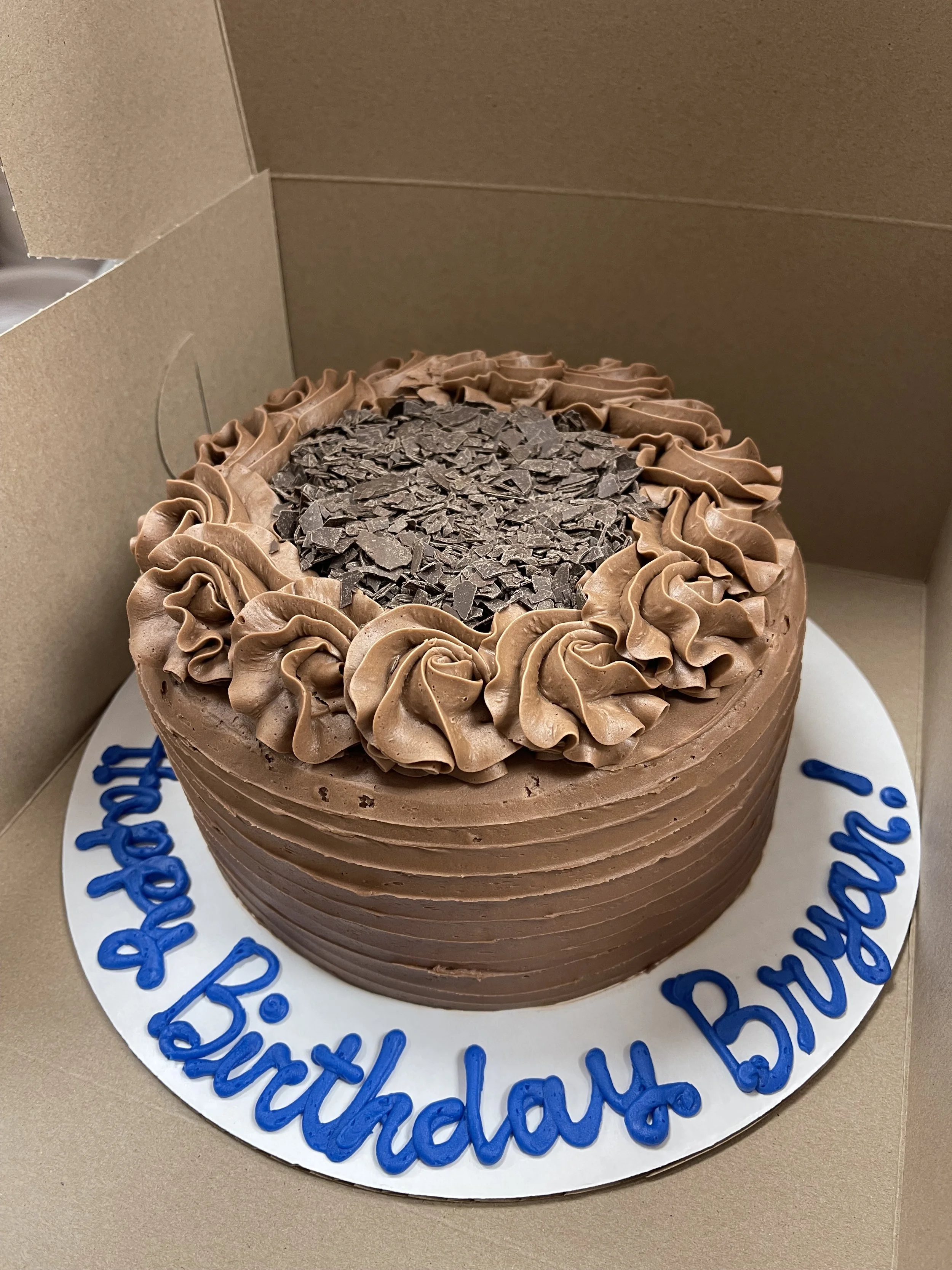 Chocolate birthday cake with chocolate frosting and chocolate shavings, inside a brown box, with blue writing on the cake board that says 'Happy 16th Birthday!'