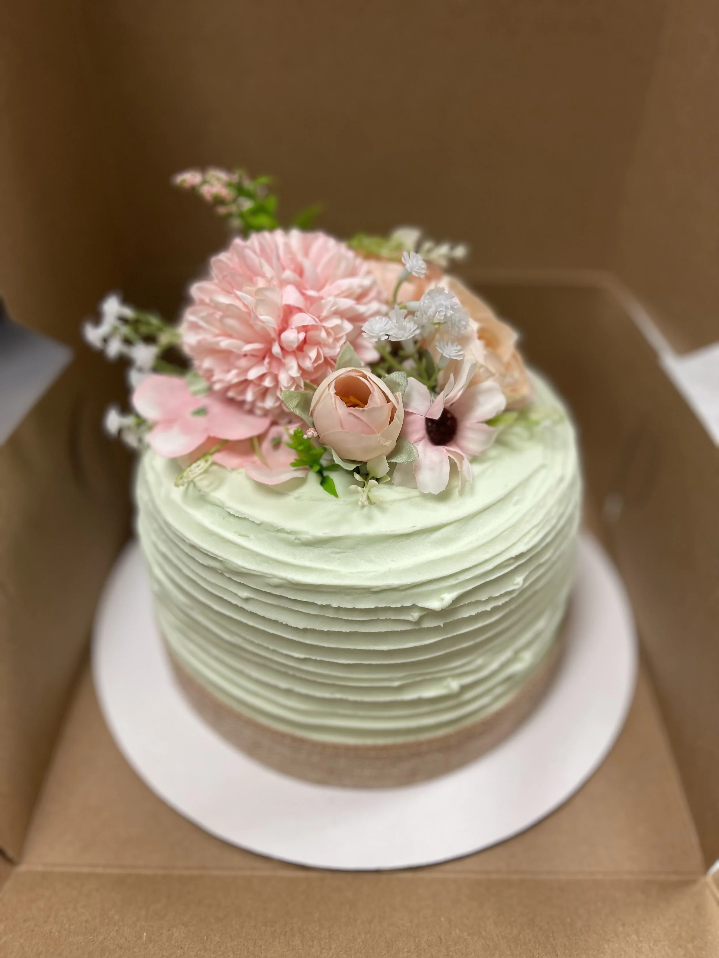 A round, layered cake with pale green frosting decorated with pink and cream-colored flowers on top, inside a cardboard box.