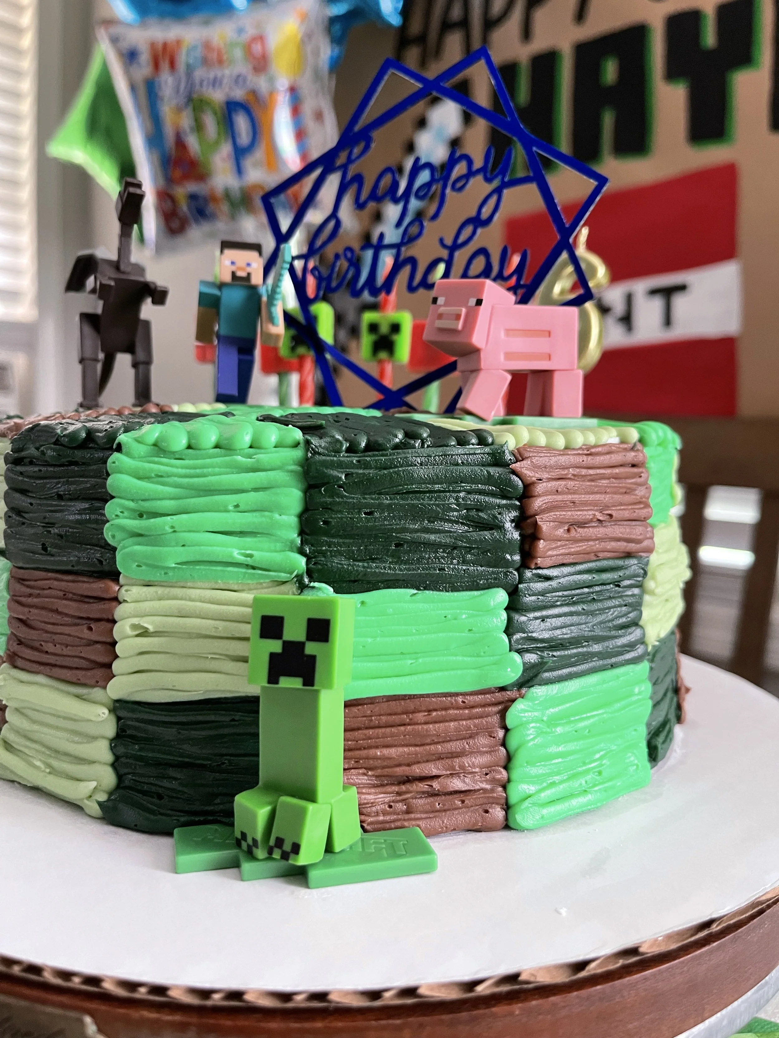 Minecraft-themed birthday cake with colorful buttercream icing blocks, decorated with figurines including a Creeper, a pig, a zombie, a skeleton, and Steve, along with a blue 'Happy Birthday' topper and balloons in the background.