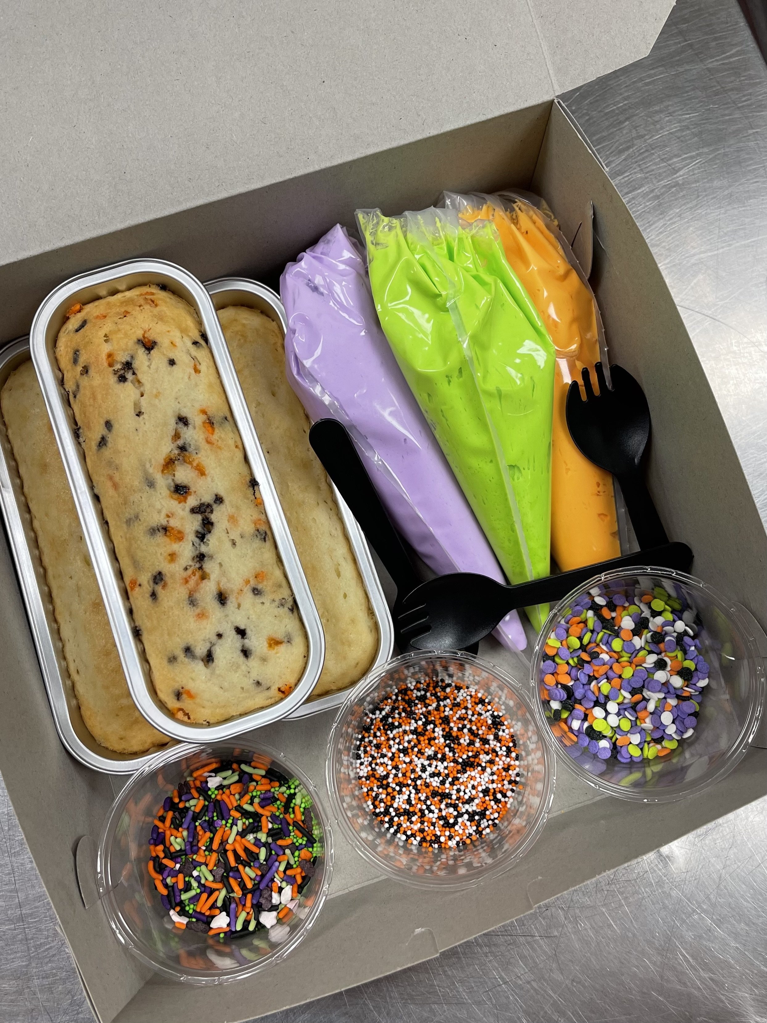 Halloween-themed cake with colored icing and various sprinkles in a box, along with black plastic forks.
