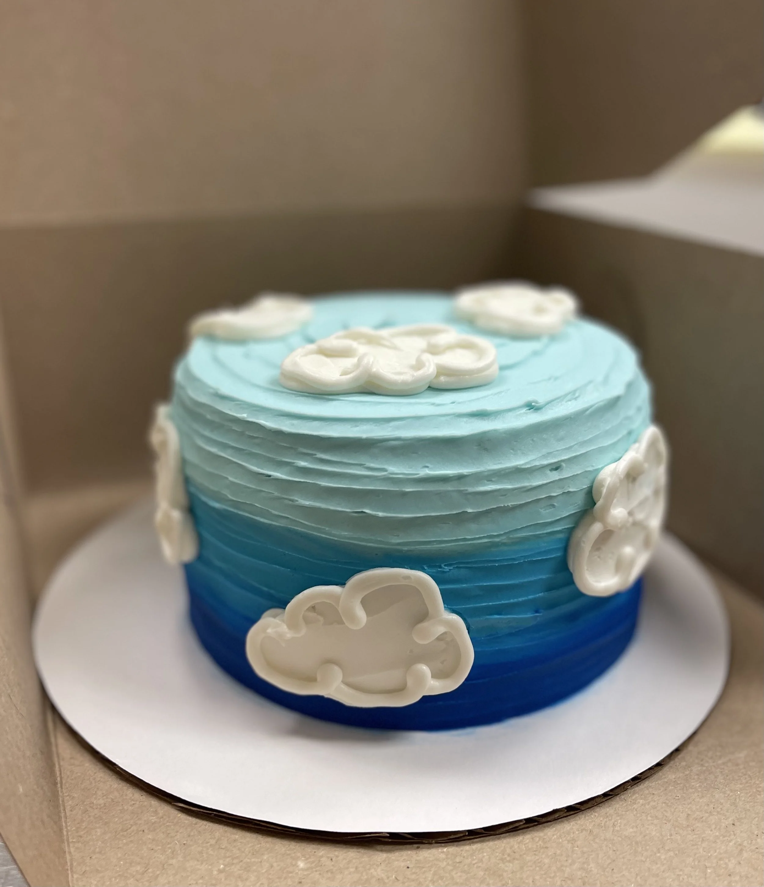 A round cake with blue ombre frosting and white cloud decorations.