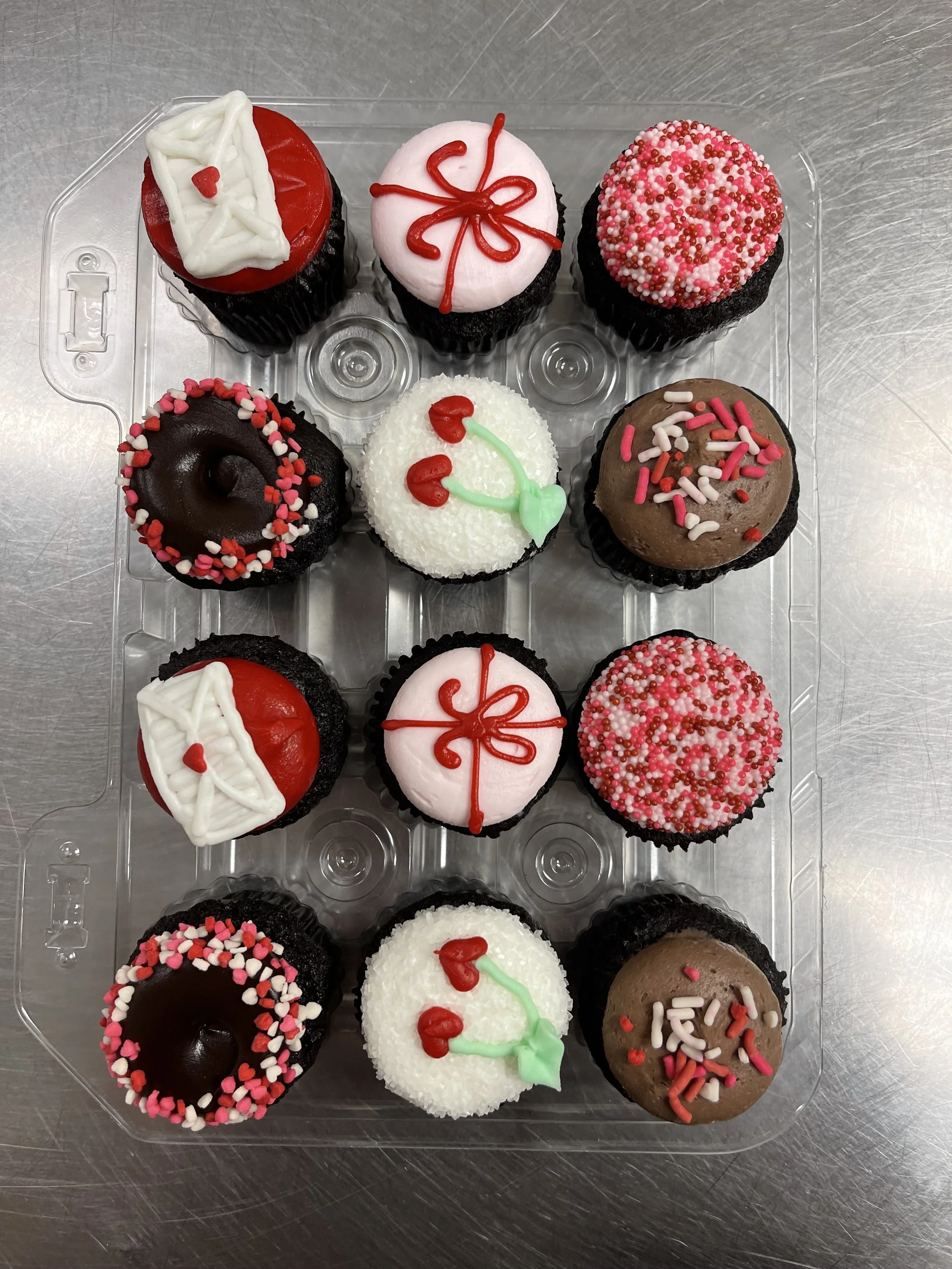 Assorted decorated cupcakes with Valentine’s Day themes include red, pink, white, and brown frosting with sprinkles, hearts, and cherries, arranged in a plastic container.