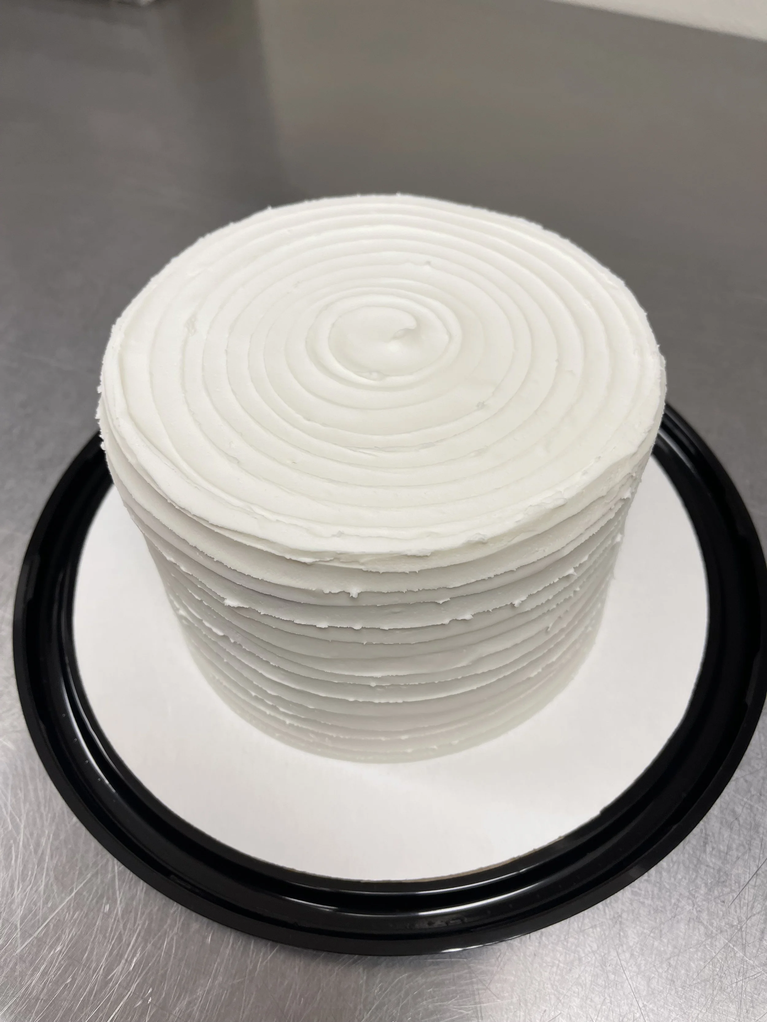 A white frosted cake with concentric circle patterns on top, placed on a round black and white cake board.