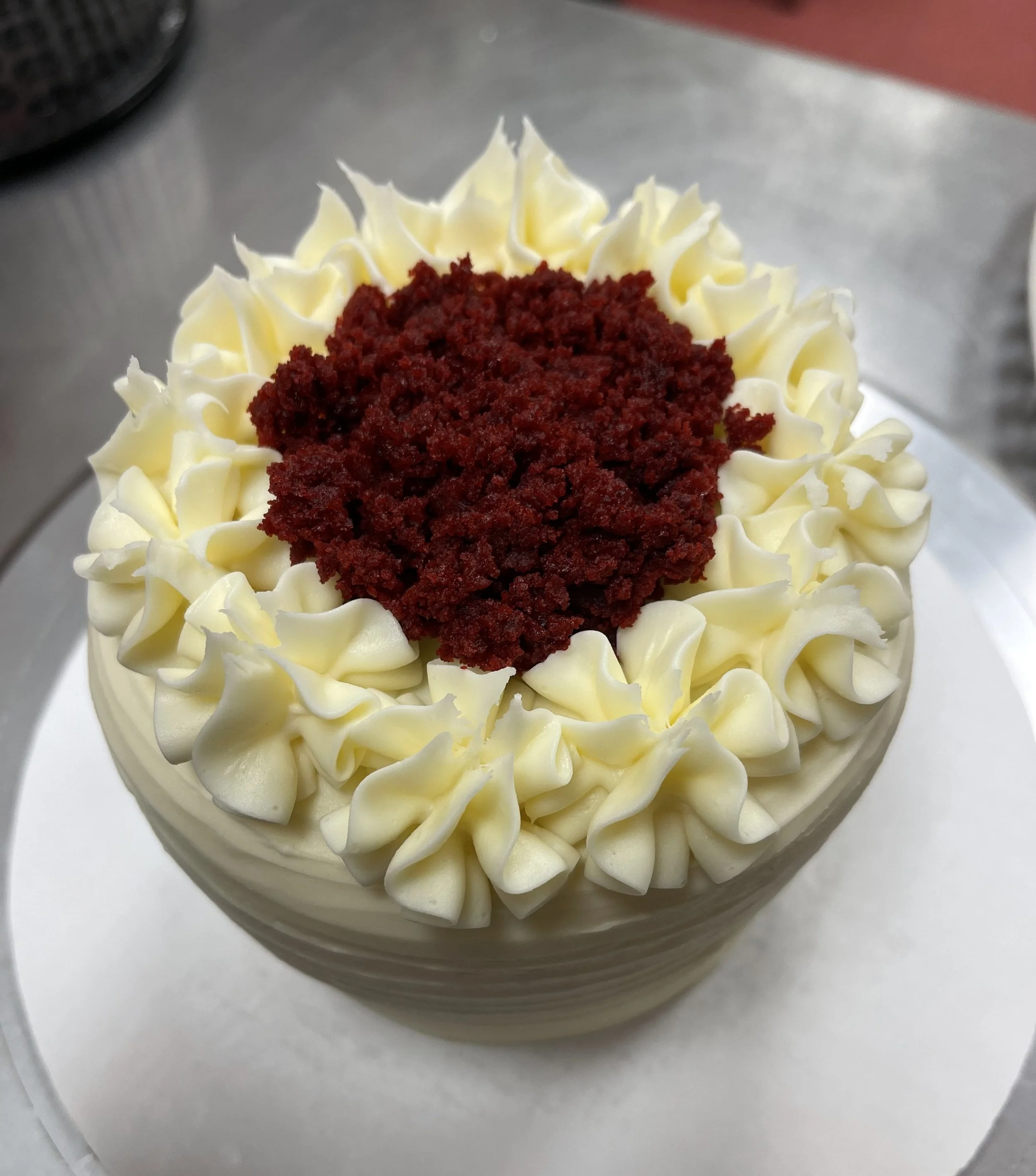 A round cake decorated with white frosting, piped in a ruffled pattern around the top edge, and topped with a red crumbly layer in the center.
