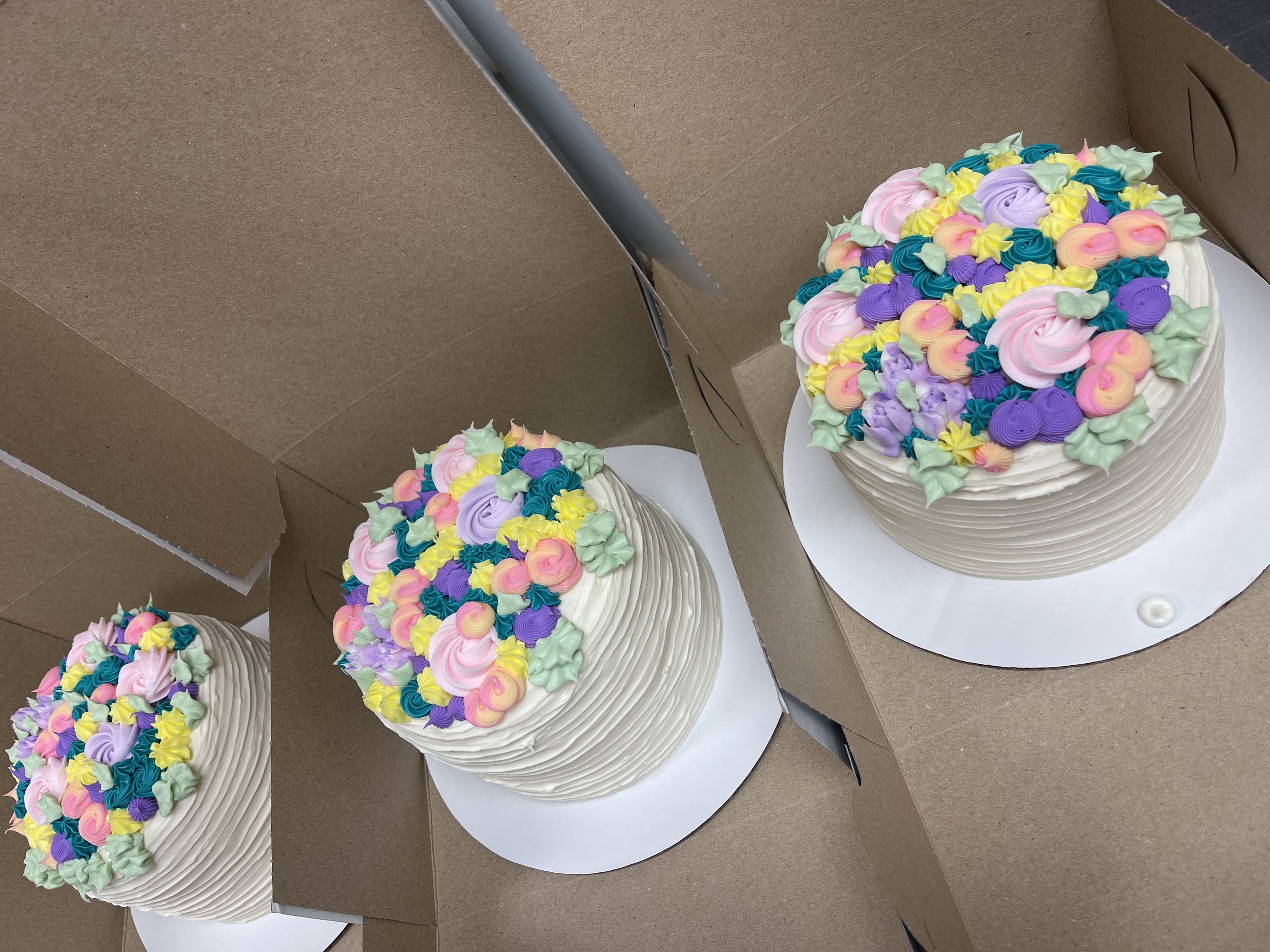Three decorated birthday cakes in open boxes, topped with colorful frosting flowers in pink, purple, yellow, blue, and green.