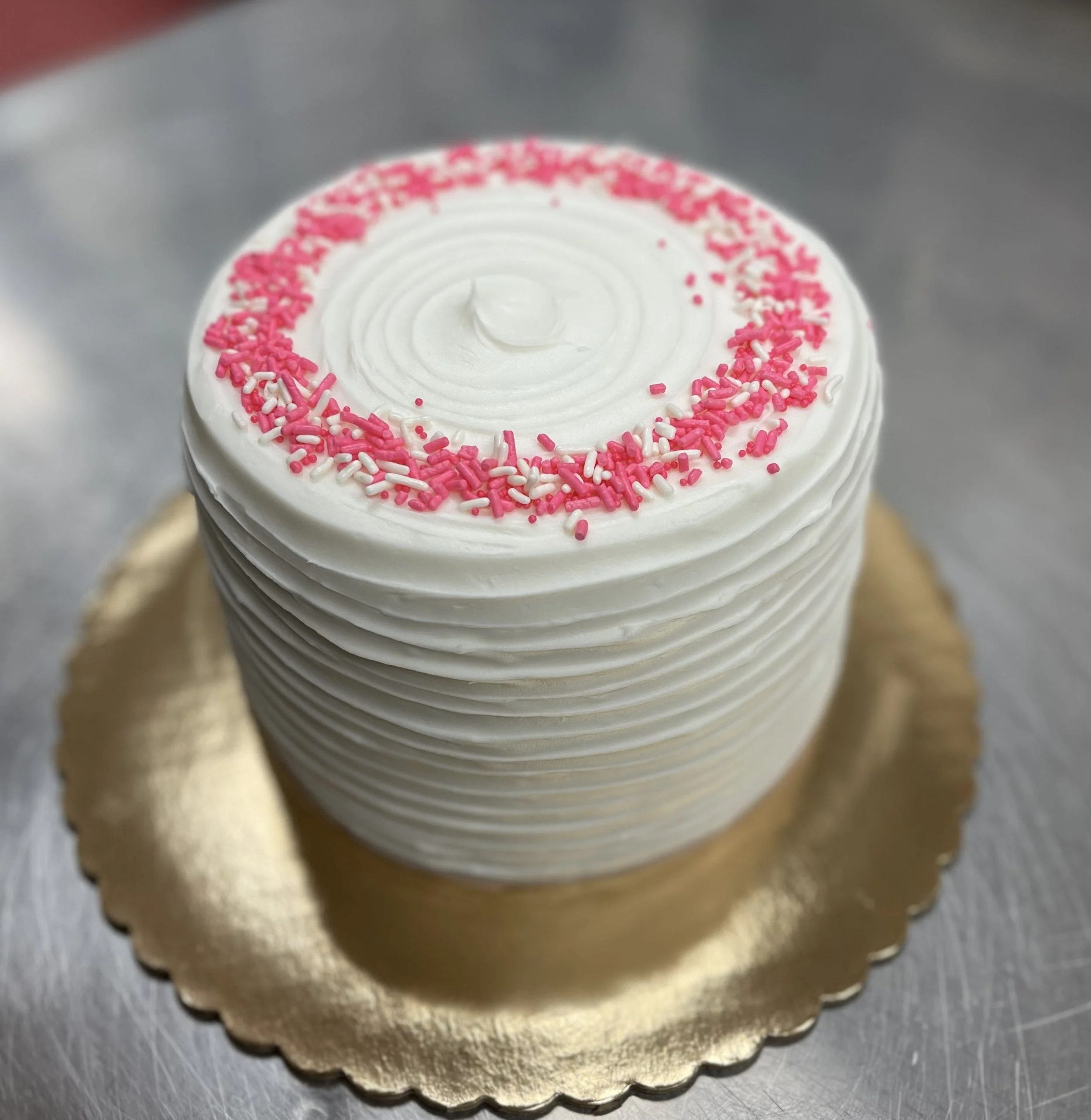 A tall, round cake with white swirl frosting, pink and white sprinkles on top, placed on a gold-colored cake board.