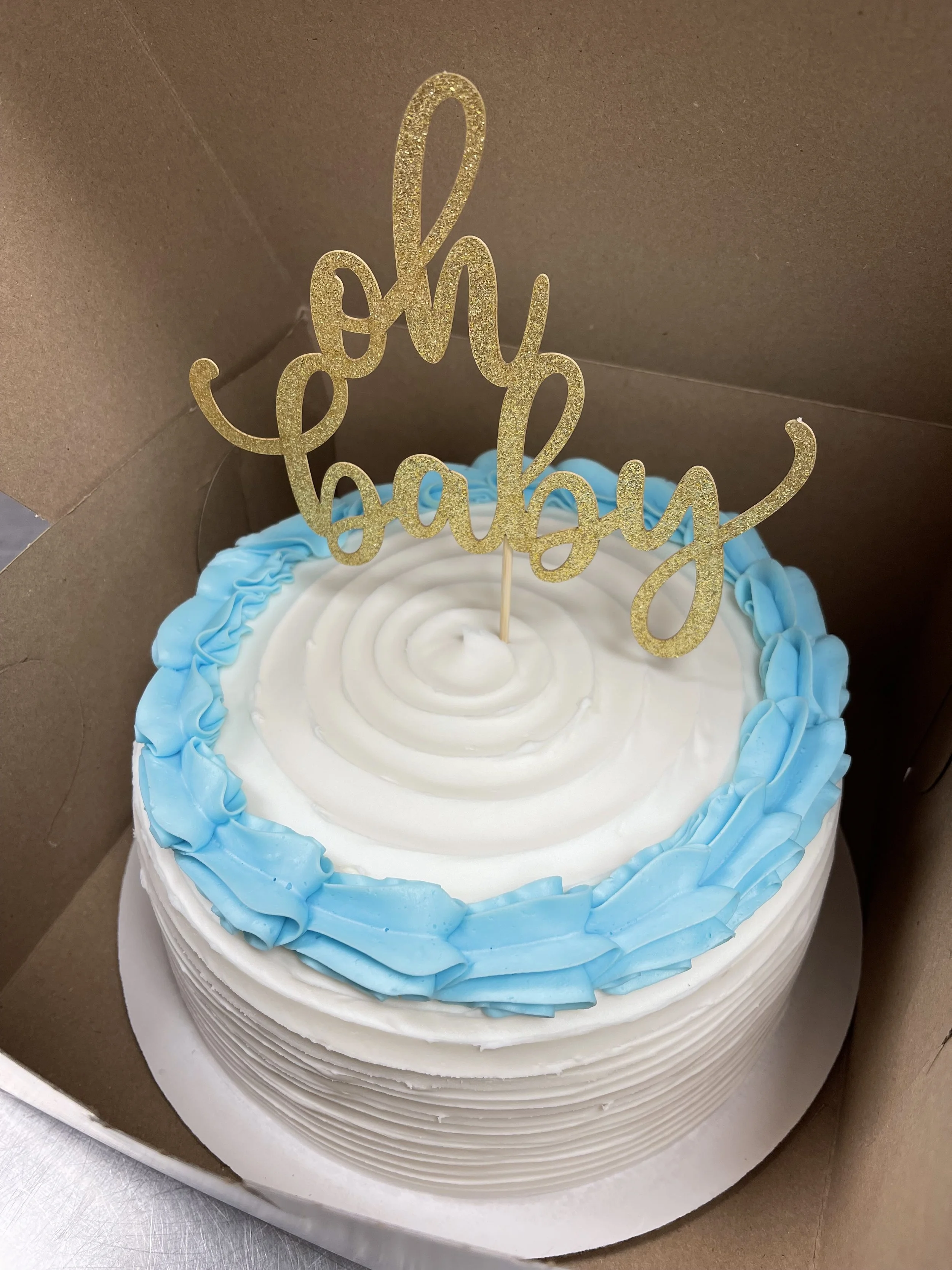 A round cake decorated with white and blue icing, with a gold glittery 'oh baby' topper, inside a cardboard box.