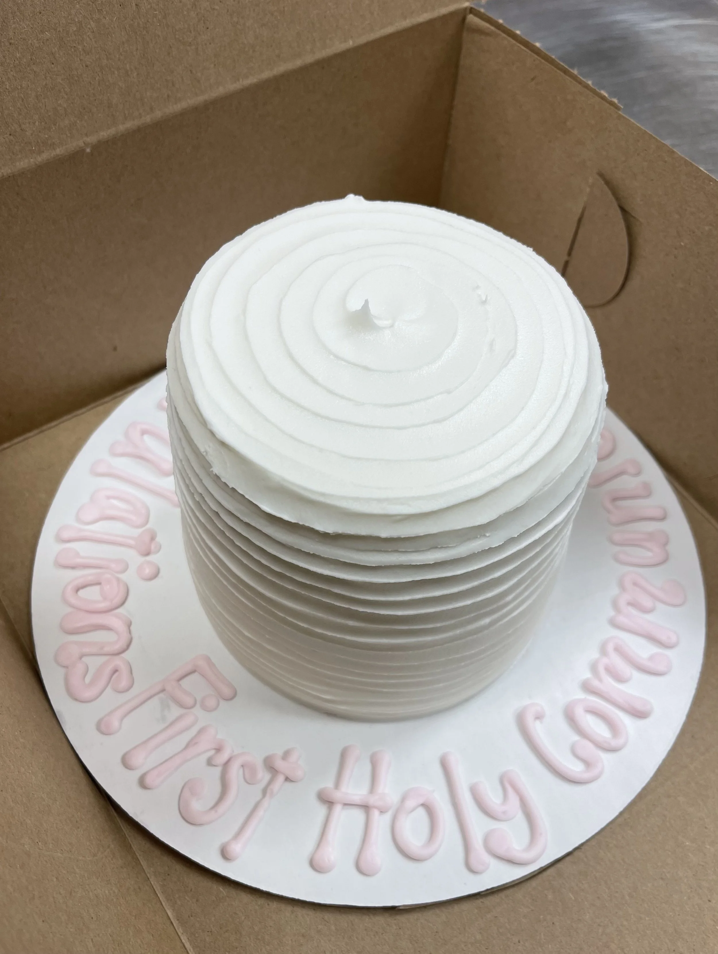 A white cake with smooth layer frosting and a spiral piped top, placed in a brown cardboard box. Pink icing around the cake reads 'Happy 4th of July'.