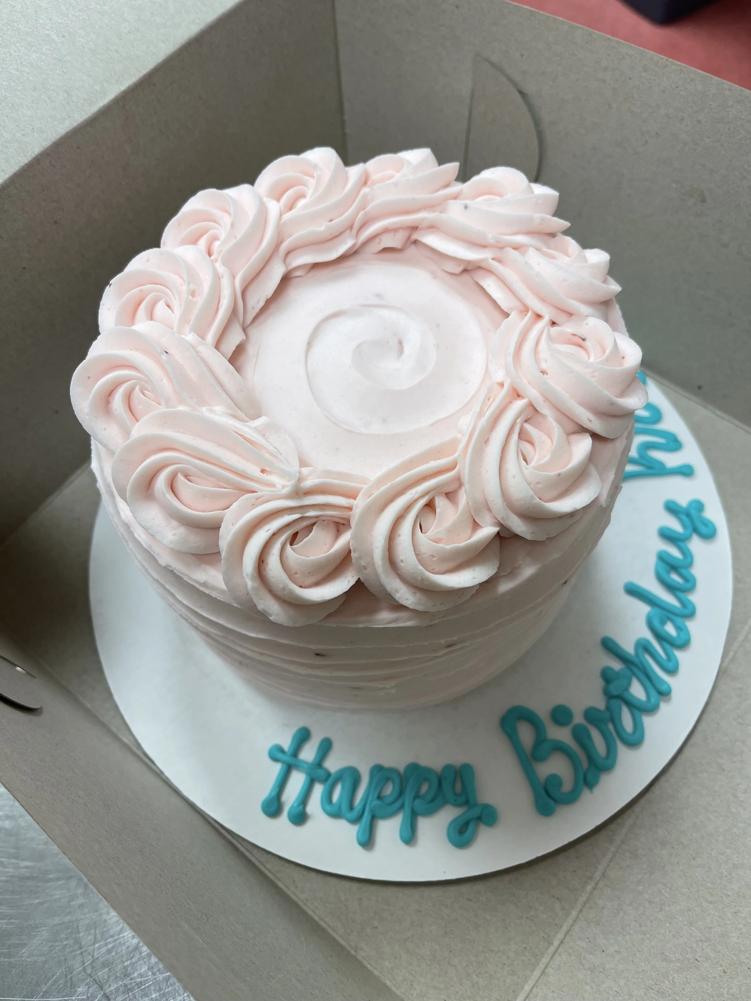A round pink birthday cake with piped frosting decorations on top, inside a cardboard box. The cake has the words 'Happy Birthday' written in blue on the white cake board.