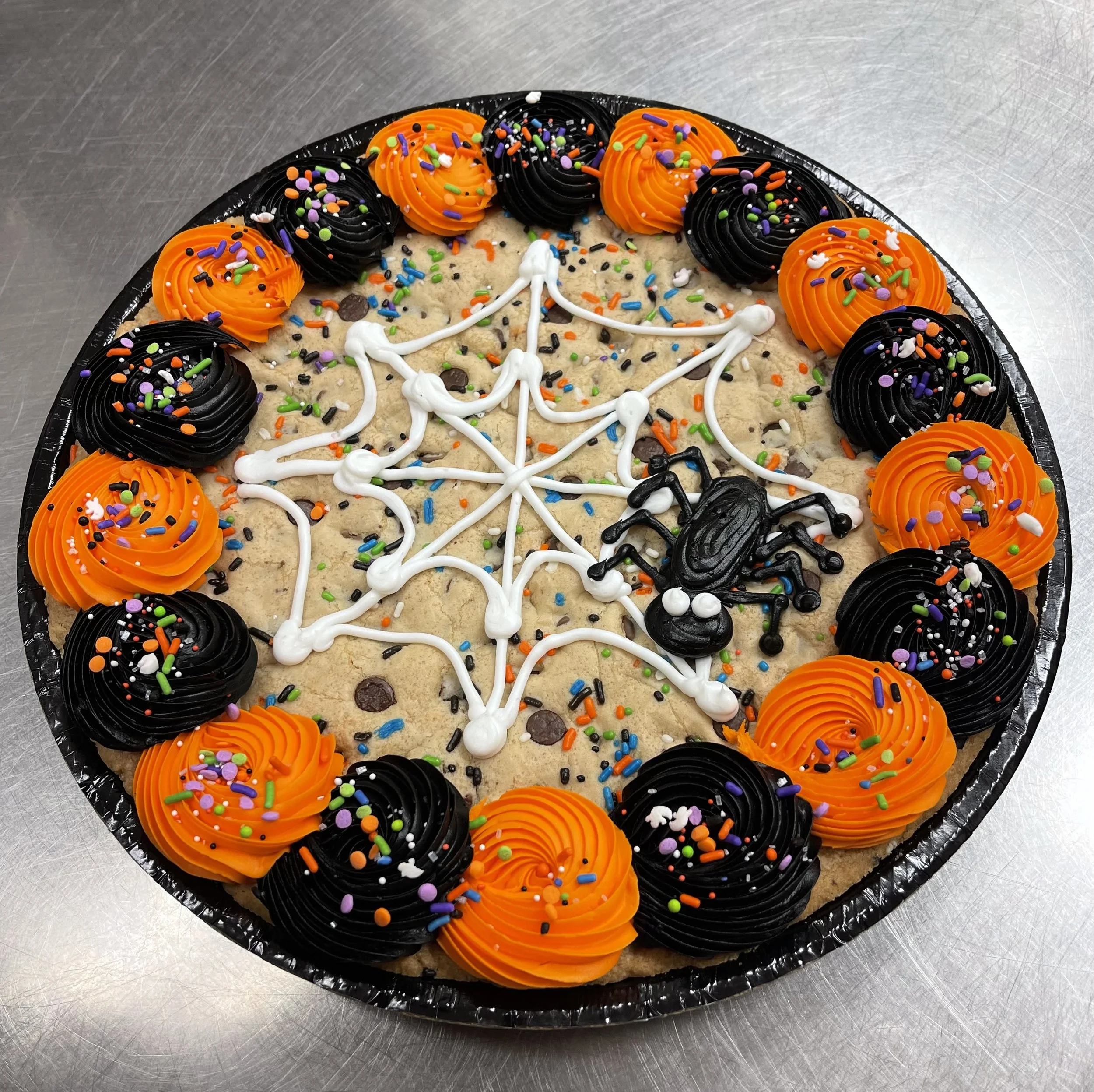 A Halloween-themed cookie cake decorated with orange and black frosting swirls, colorful sprinkles, and a spider with a web drawn in white icing.