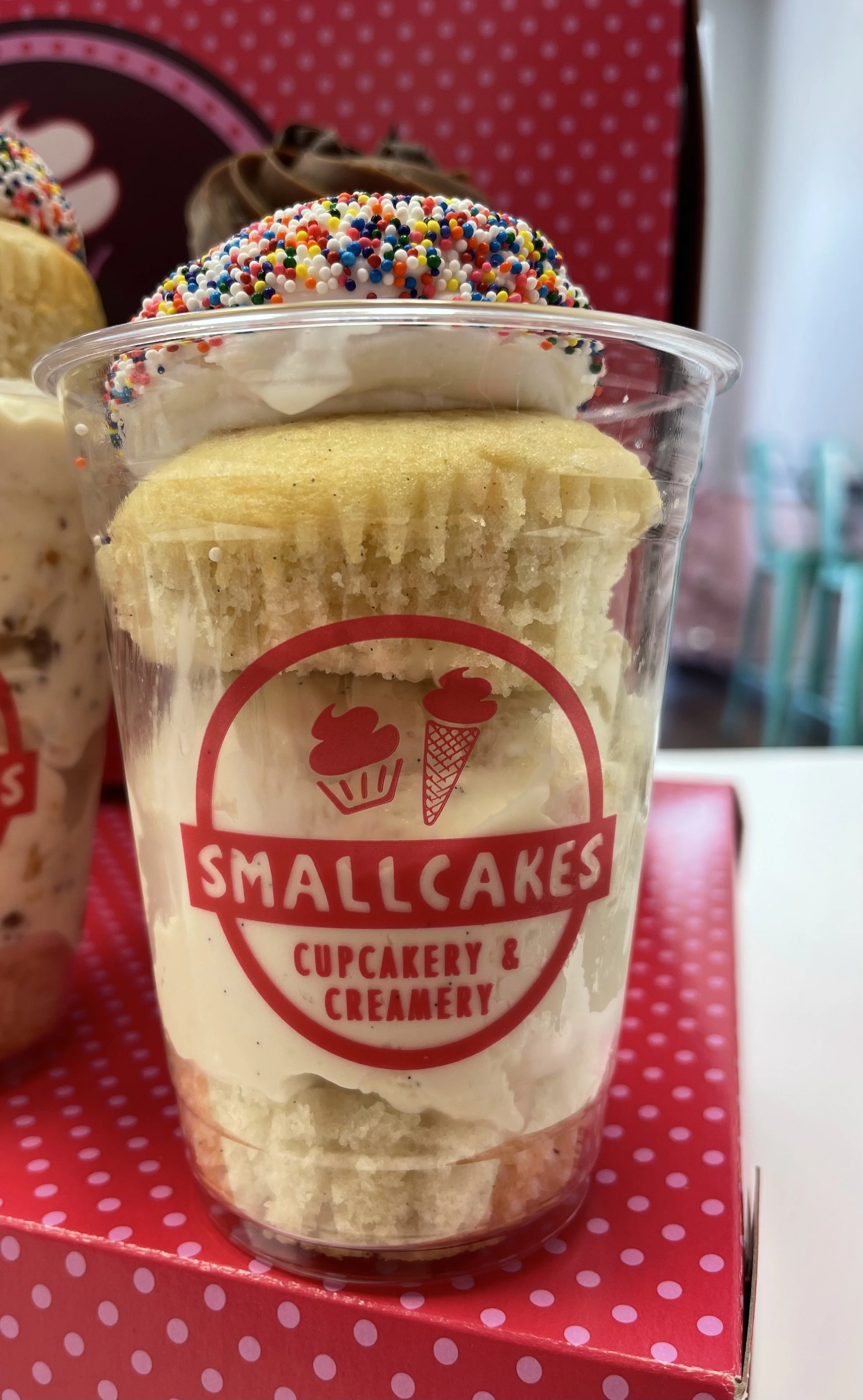 Cupcake in a clear plastic cup with white frosting, rainbow sprinkles, and a yellow cake layer, labeled 'Smallcakes Cupcakery & Creamery' with a red logo of a cupcake and ice cream cone.