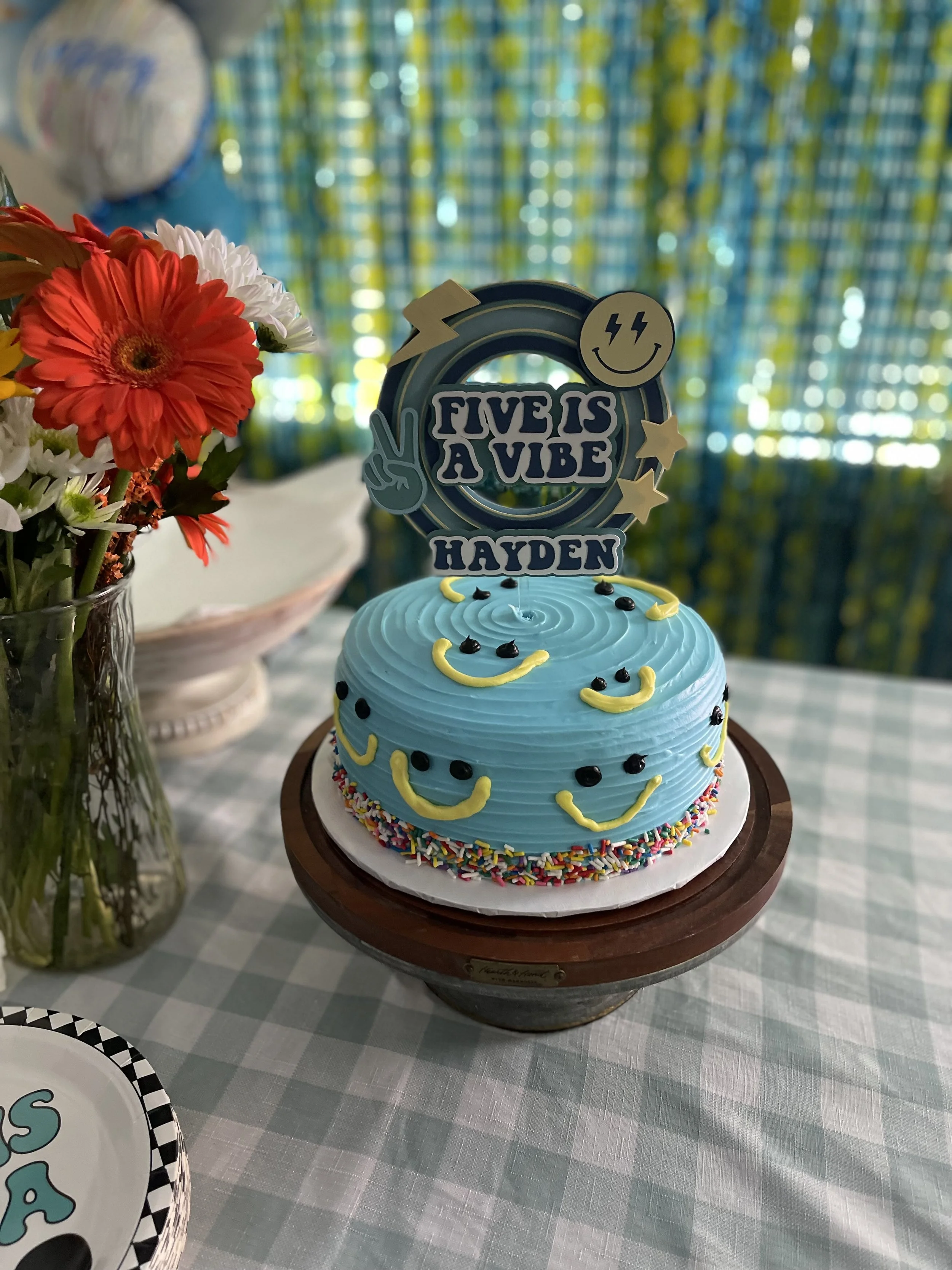 A birthday cake with blue frosting decorated with yellow and black smiley faces, colorful sprinkles around the base, and a cake topper that reads "FIVES A VIBE" with supplemental graphics and the name "HAYDEN" on it. The cake is on a wooden cake stan