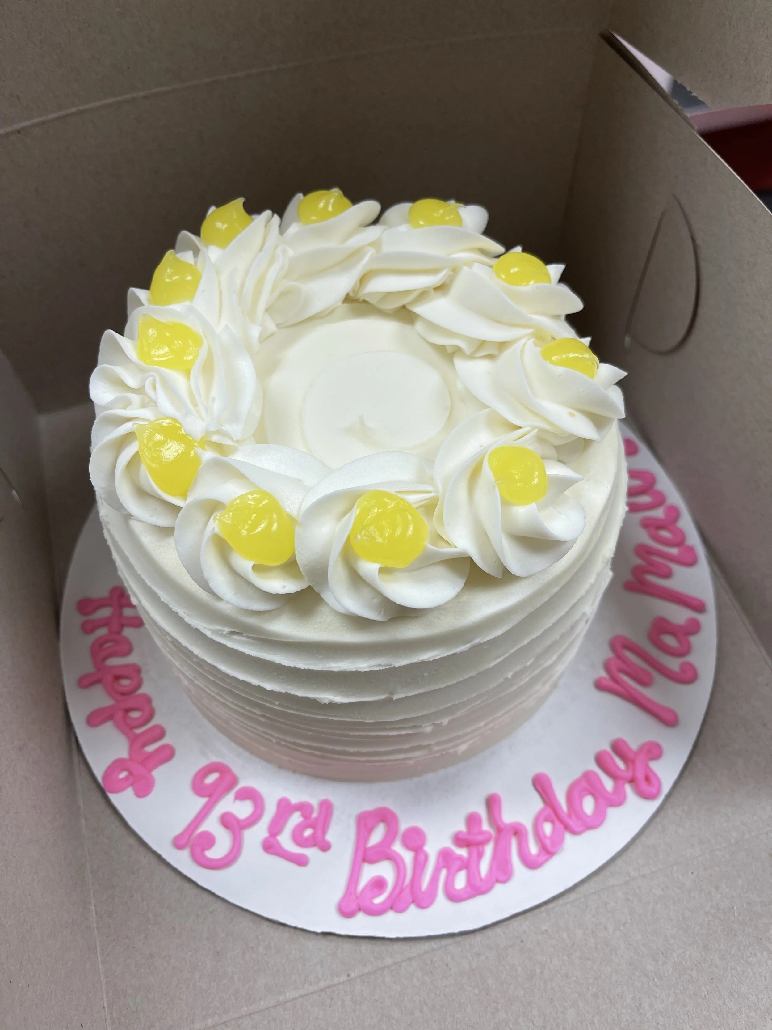 A birthday cake decorated with white frosting, yellow cherries, and pink writing on the base that reads 'Happy 13th Birthday Maria/Mam'