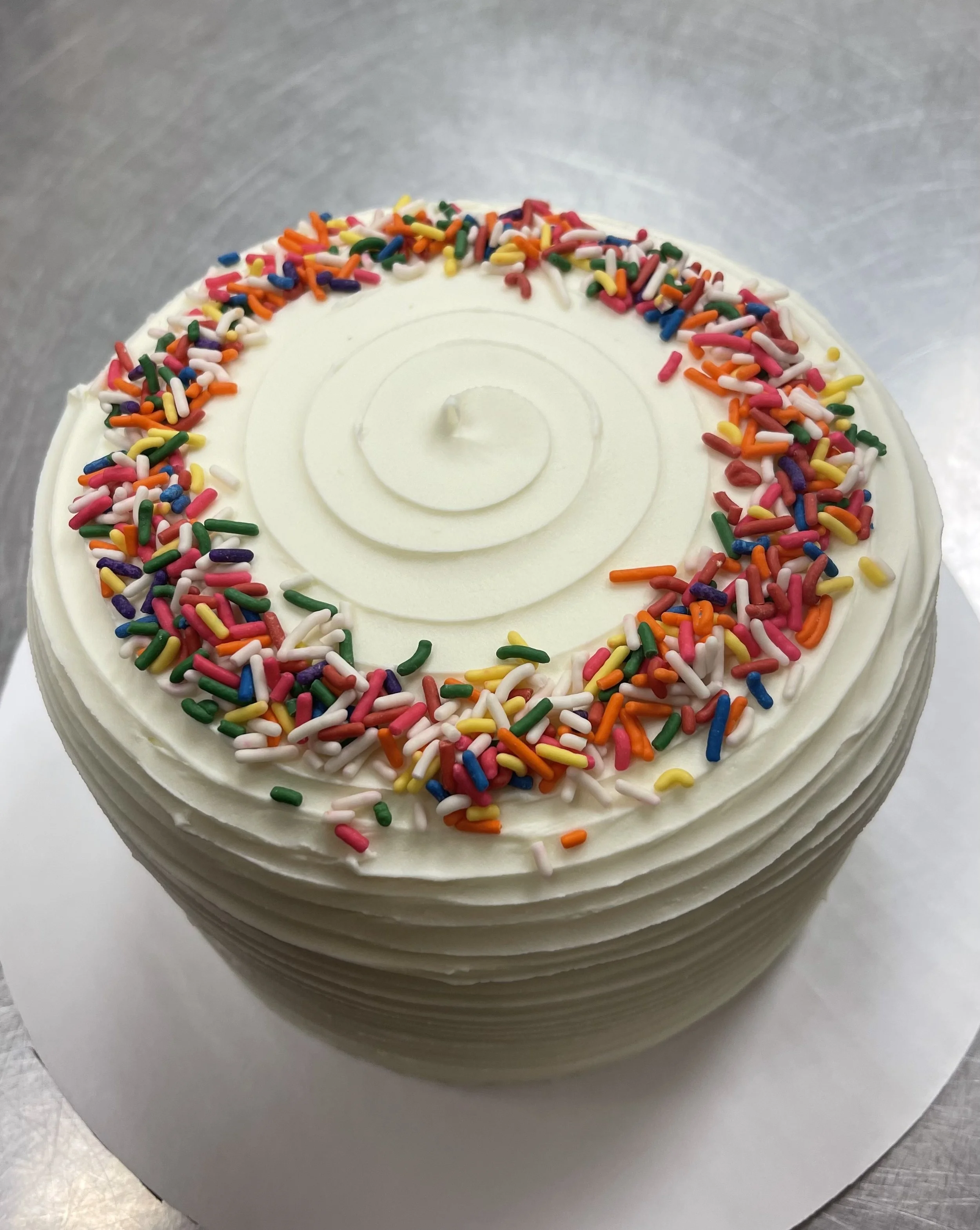 A round cake with white icing, colorful rainbow sprinkles, and a white spiral decoration on top, on a white surface.