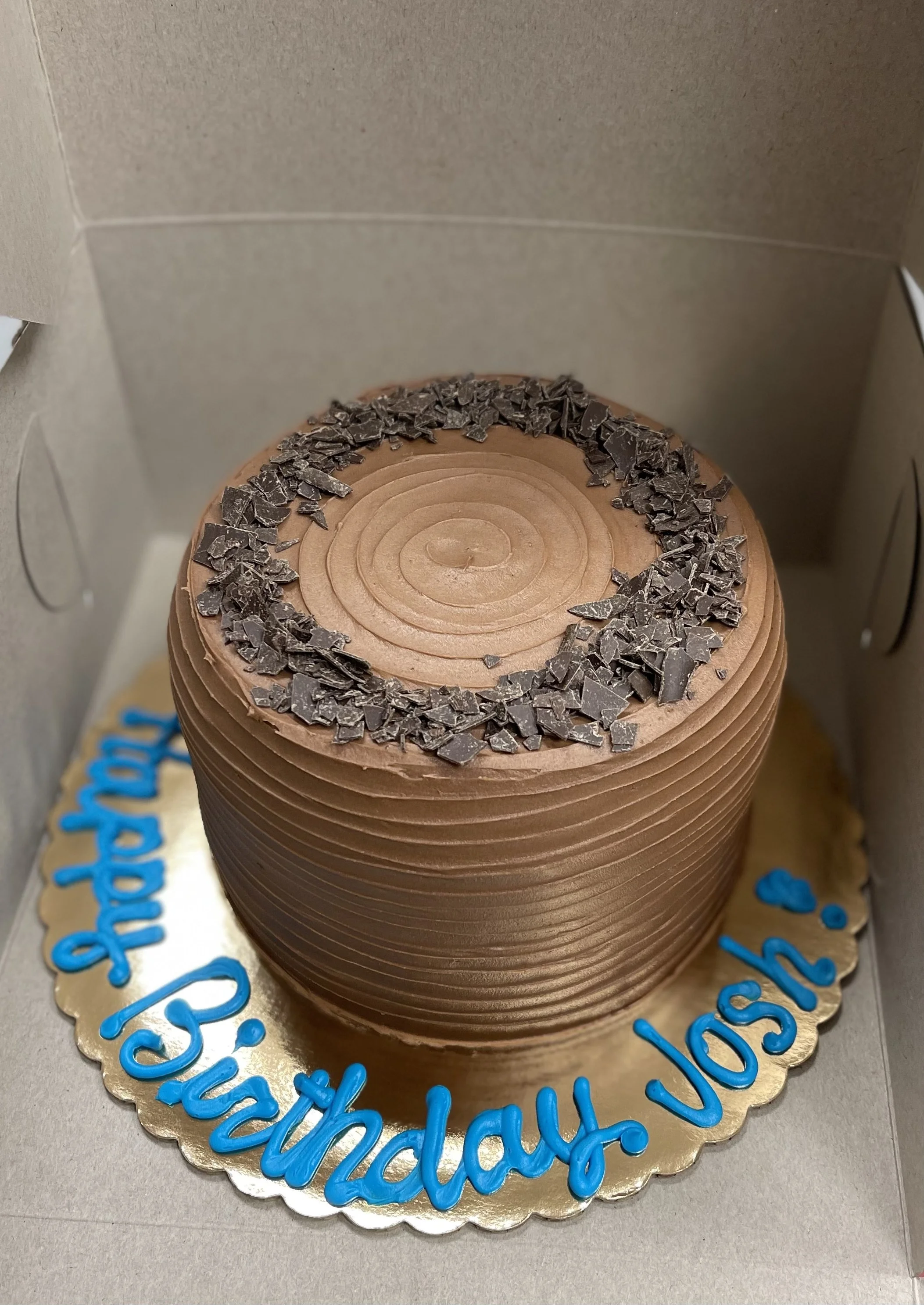 A chocolate birthday cake in a box with chocolate shavings on top and a message that reads "Happy 18th Birthday Joshua" written in blue icing.