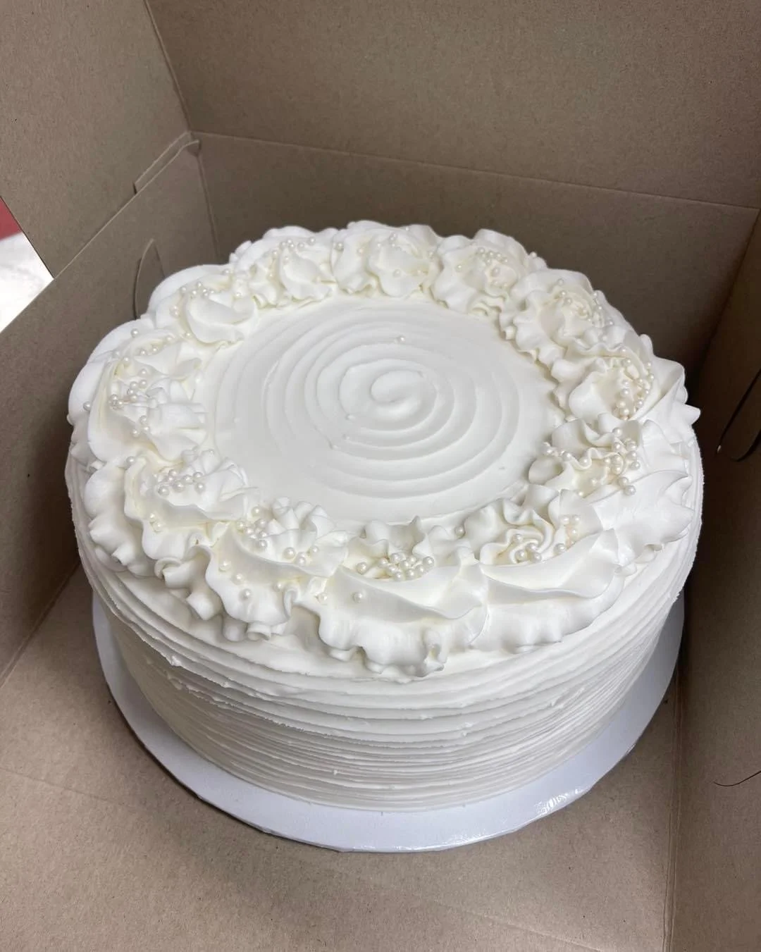 A round white cake with decorative icing, pearls, and ruffled icing border, inside a cardboard box.