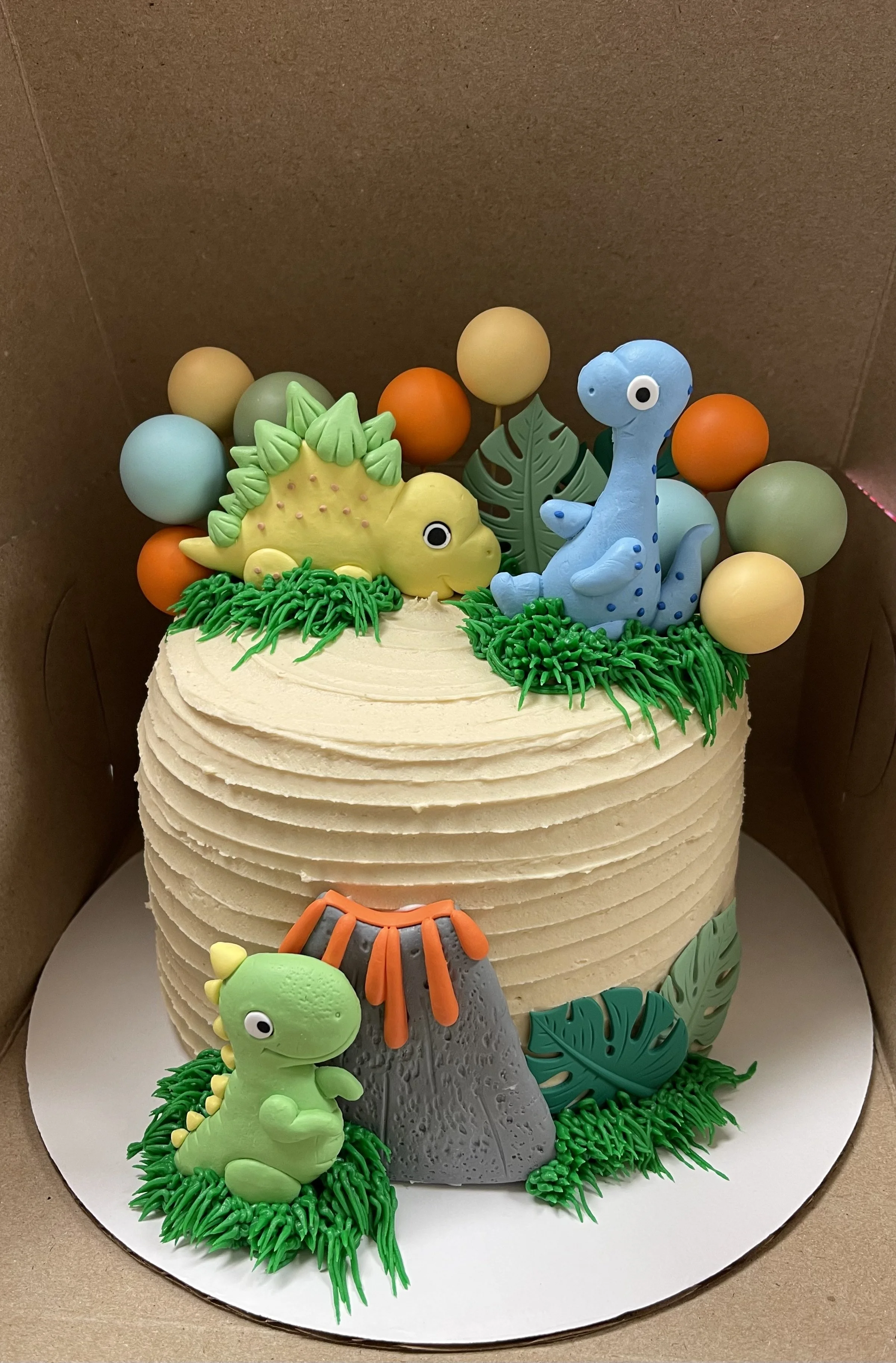A children's birthday cake decorated with colorful dinosaur-themed fondant figures, including a green stegosaurus, a blue sauropod, and a green baby dinosaur, surrounded by green grass-like icing, decorative balls, and leaves.