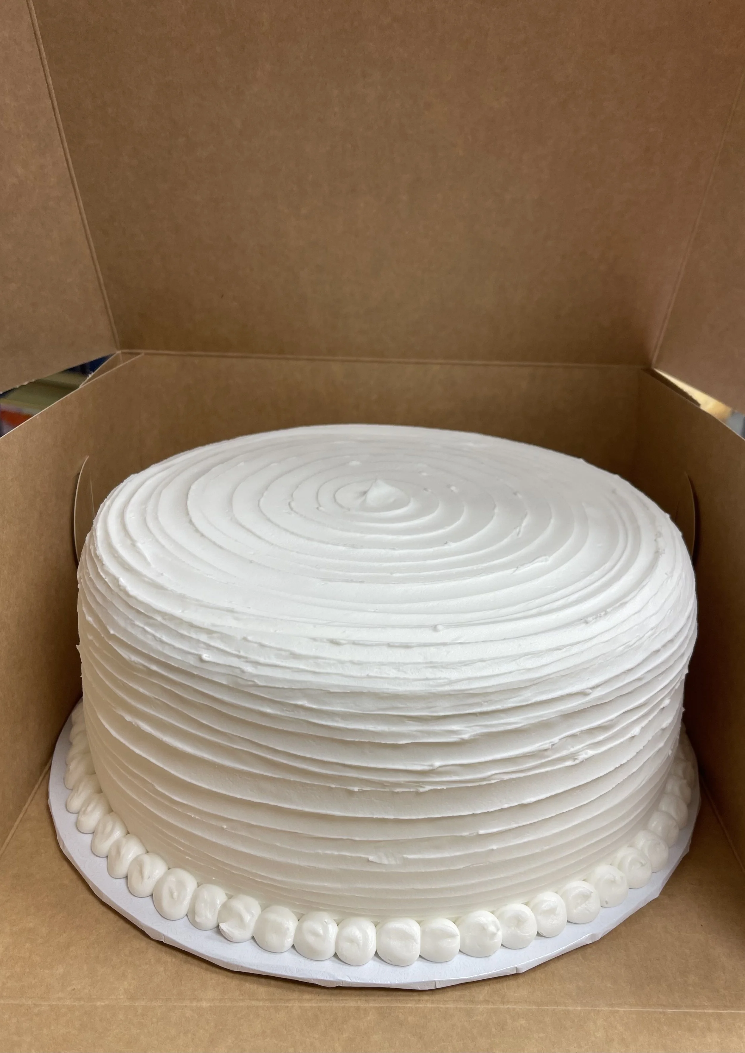 White frosted round cake with a swirl design, sitting in a cardboard box.