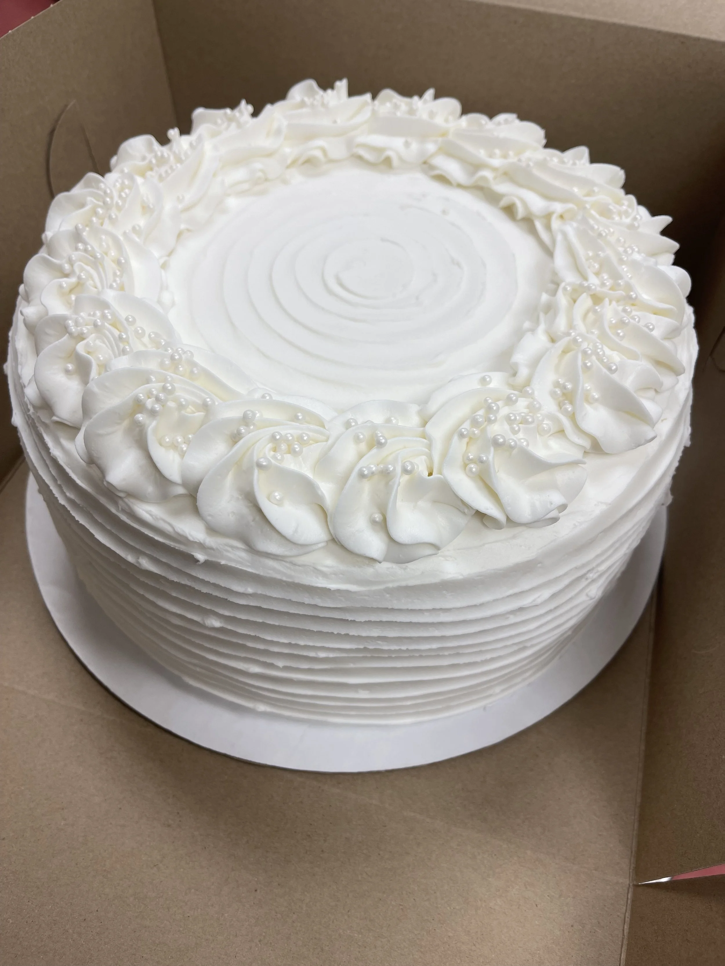 A round white cake with piped whipped cream decoration around the top edge, decorated with small edible pearls, in a brown cardboard box.