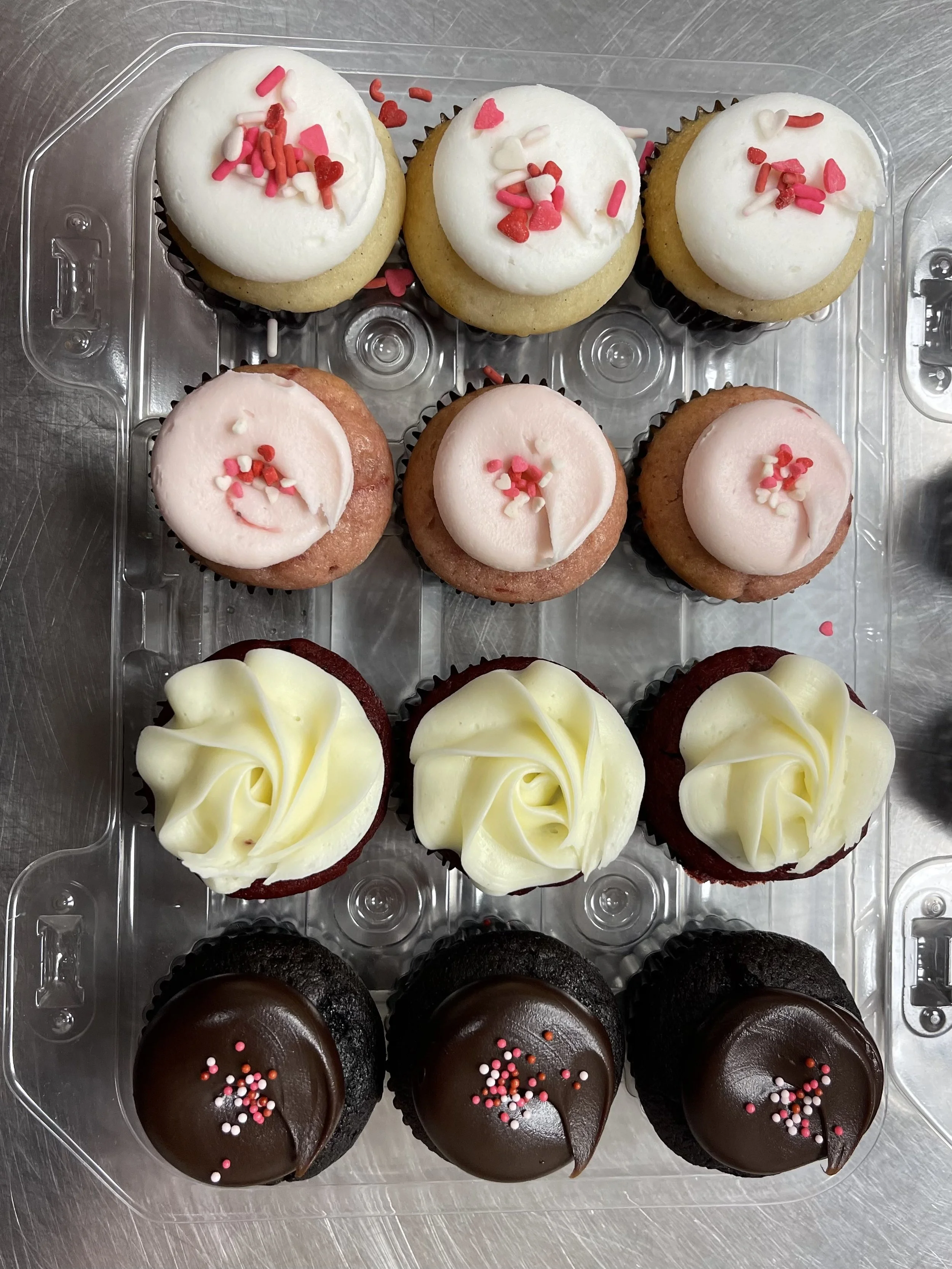 A tray of nine decorated cupcakes with white, pink, and chocolate frosting, some topped with sprinkles, arranged in rows of three.
