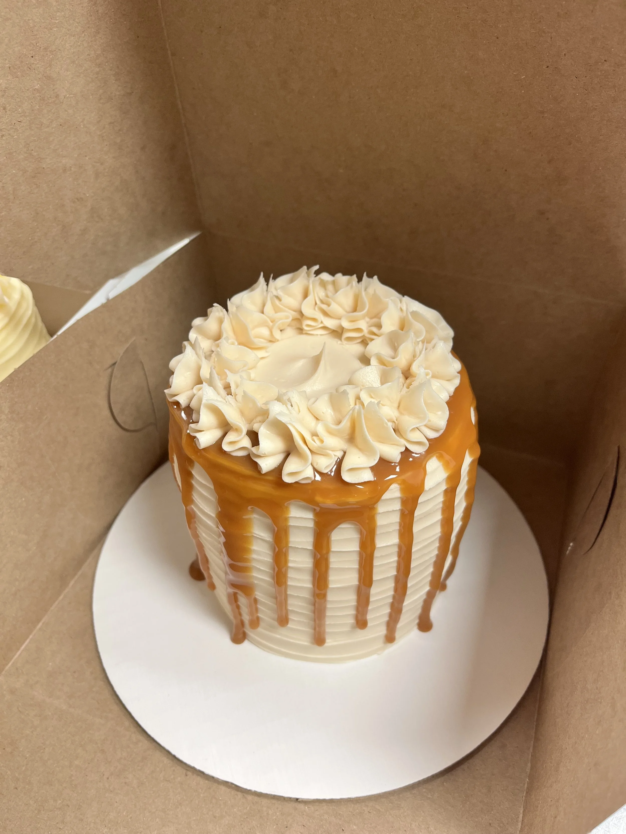 A tall birthday cake with white frosting, caramel drizzle, and decorative white icing flowers on top in a brown cake box.