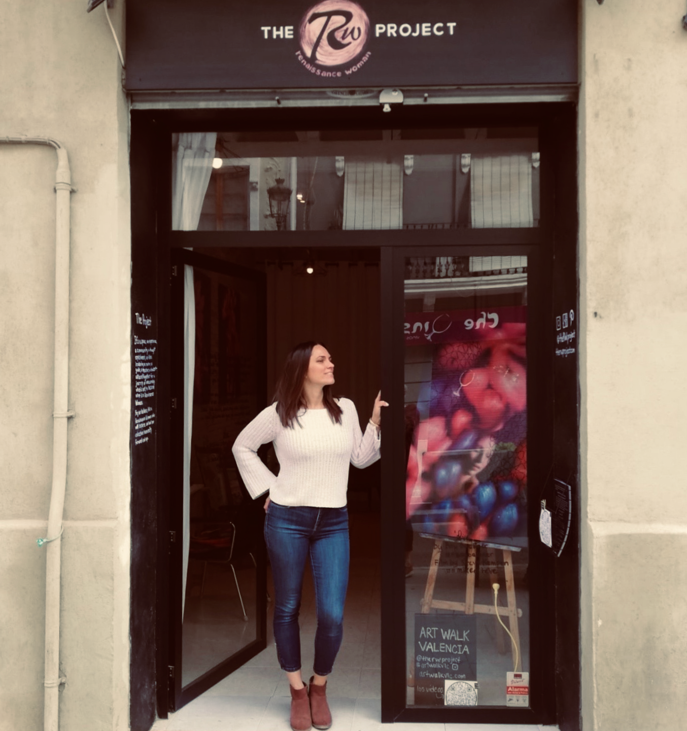 A woman with dark hair wearing a white sweater and blue jeans standing at the entrance of a store named 'The Revival Project.' She is touching the door and smiling. The store has a sign that reads 'Art Walk Valencia' and features an art display with colorful artwork visible inside.