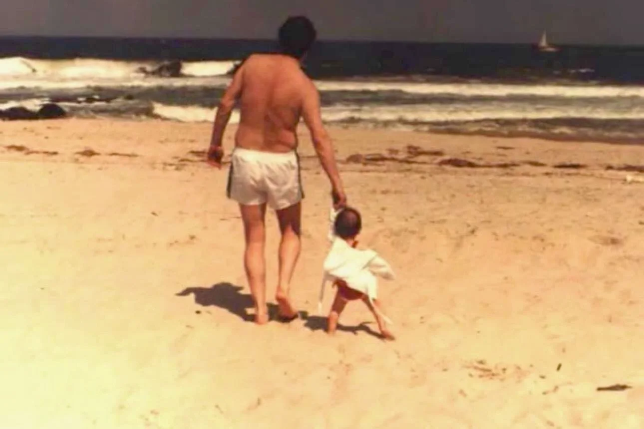 A man and a small child walking hand in hand on a sandy beach.