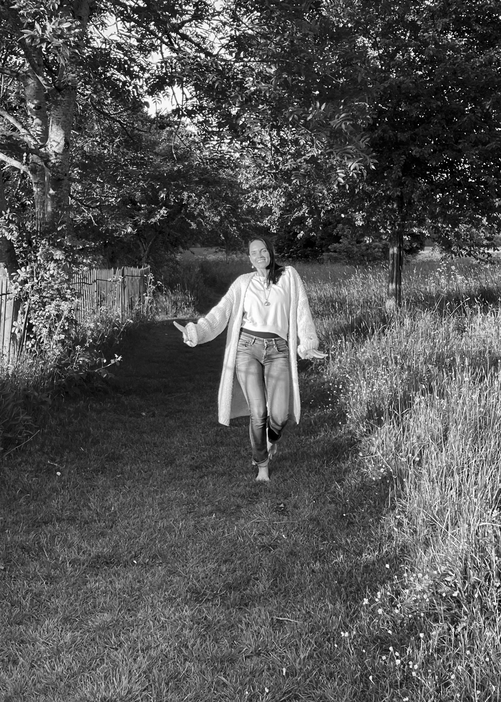 A woman smiling and walking barefoot on a grassy path in a park, surrounded by trees and vegetation.