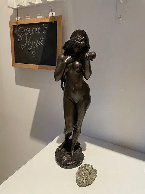 Bronze sculpture of a nude woman with long hair, covering her face and breasts, with a chalkboard in the background that reads "Gracie's Mox"