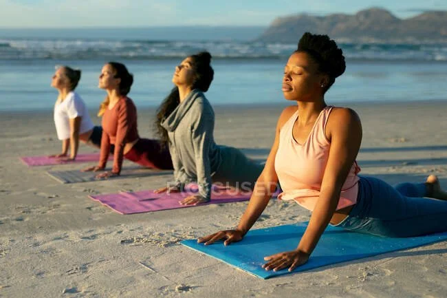 Four women practicing yoga on mats on the beach with ocean waves and distant cliffs in the background.