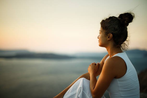 A woman sitting on a beach during sunset, looking out towards the horizon.