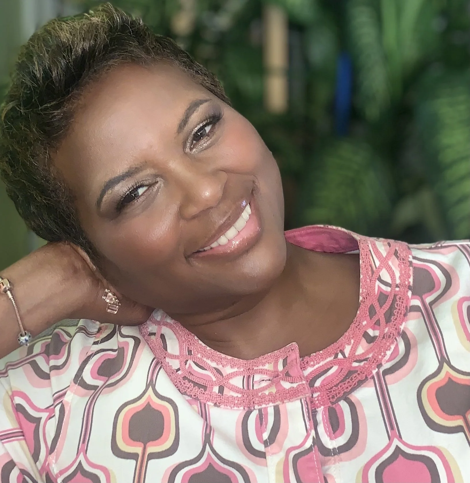 Close-up of a smiling woman with short dark hair, wearing a colorful patterned top with pink lace trim and jewelry, set against a green leafy background.