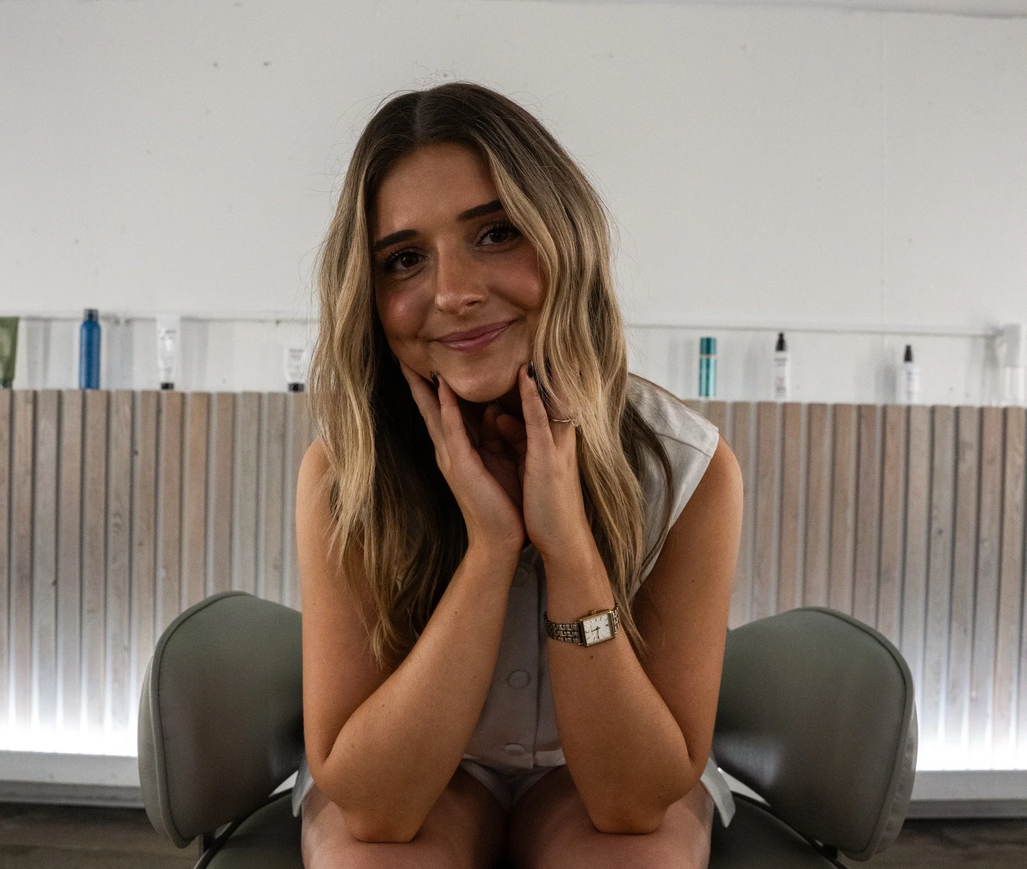 A young woman with long wavy blond hair smiling at the camera while sitting on a gray chair in an indoor setting with a light-colored wall and a wooden textured background.