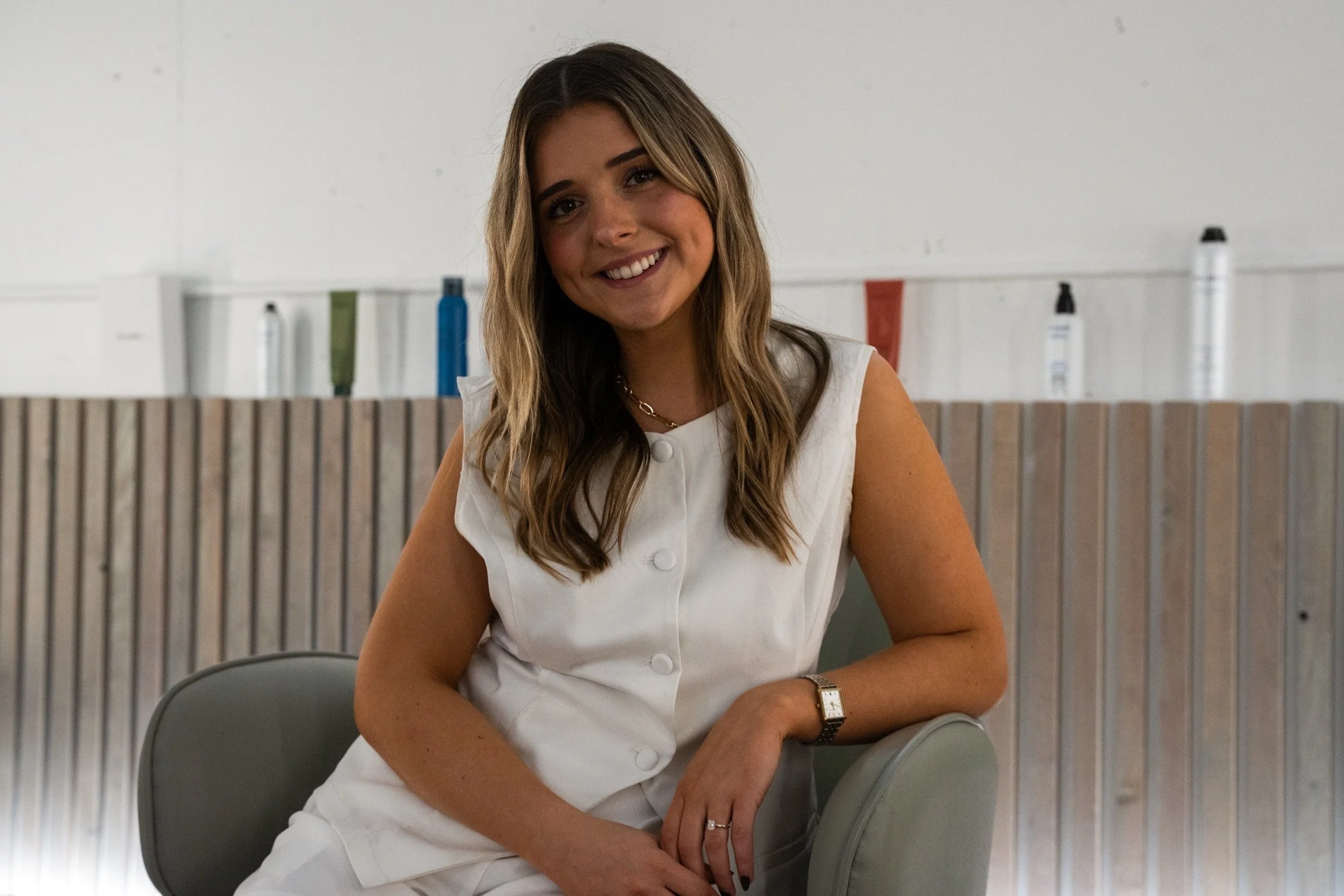 A smiling woman with long wavy brown hair sitting on a gray chair, wearing a sleeveless white dress and a gold watch, in a modern indoor setting.