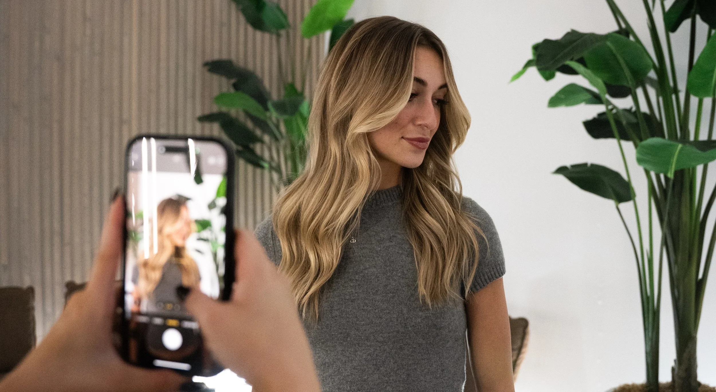 A woman with long wavy blonde hair posing for a photo indoors with large green plants in the background, while someone takes her picture with a smartphone.