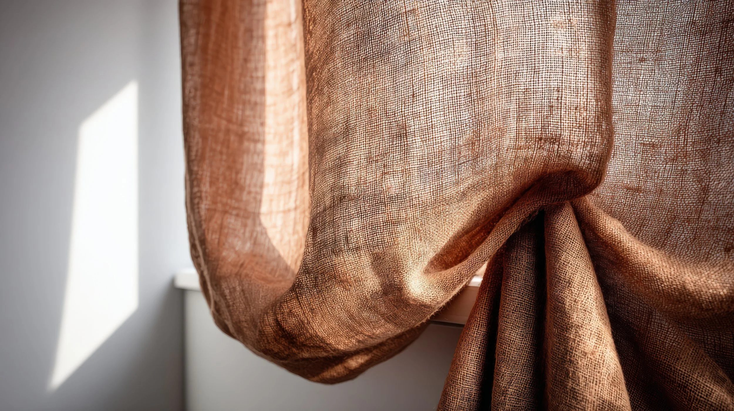 Close-up of a sheer, brownish, textured curtain tied in a knot, with sunlight casting shadows on a white wall in the background.