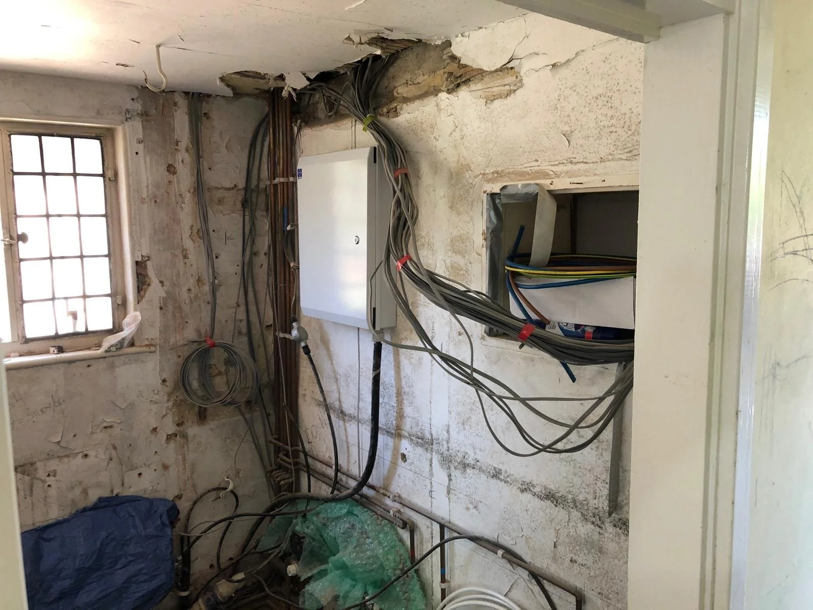 Electrical wiring and a small electrical box on an unfinished wall with exposed pipes and a window.