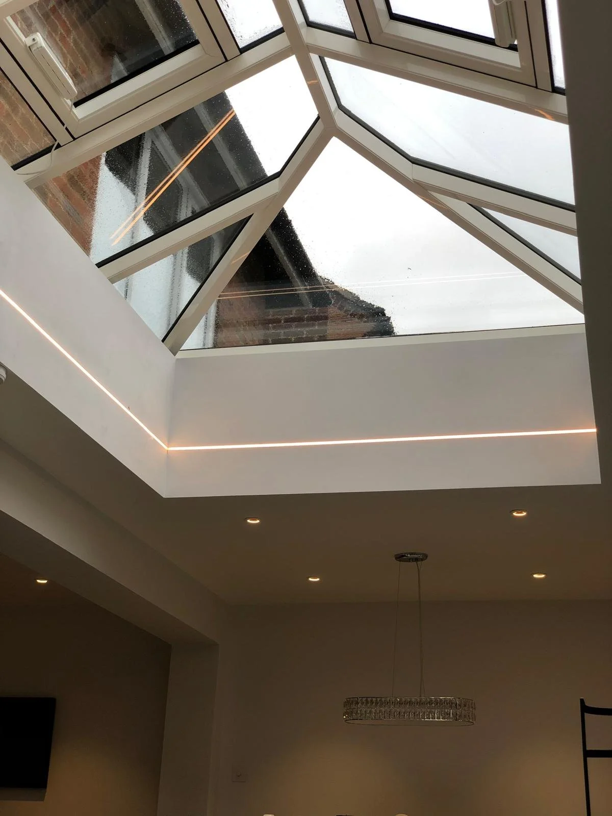 Interior view of a modern room with a large glass skylight ceiling, rain on the glass, white walls, recessed ceiling lights, and a decorative hanging light fixture.