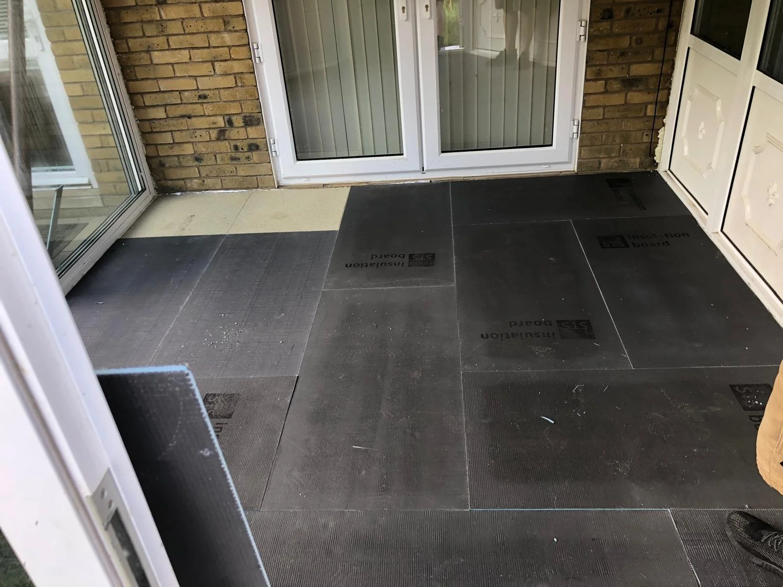 View of a covered porch or patio area with black insulation boards on the floor, a glass sliding door, and a white door with decorative details. There is a brick wall on the left side.