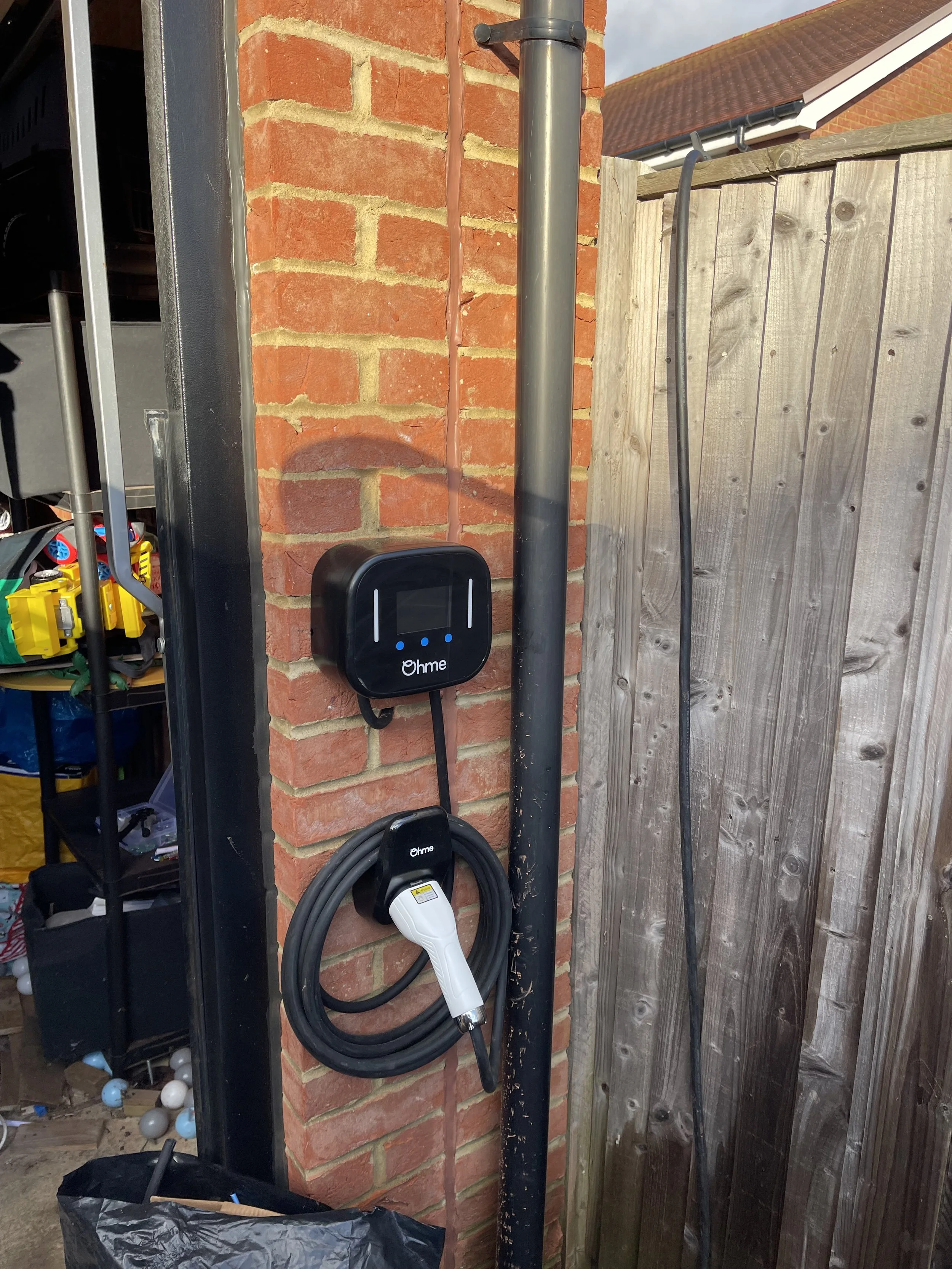 An electric vehicle charging station mounted on a brick wall with a black and white charging cable hanging below it, next to a wooden fence.