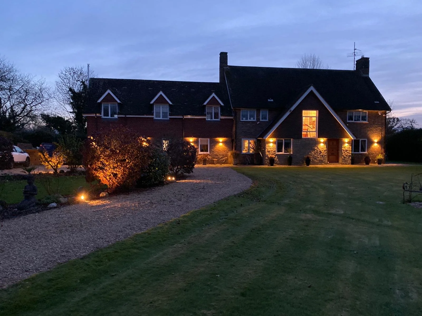 A large house at dusk with outdoor lighting illuminating the exterior, surrounded by a well-maintained lawn and garden, with a gravel pathway leading up to the entrance.