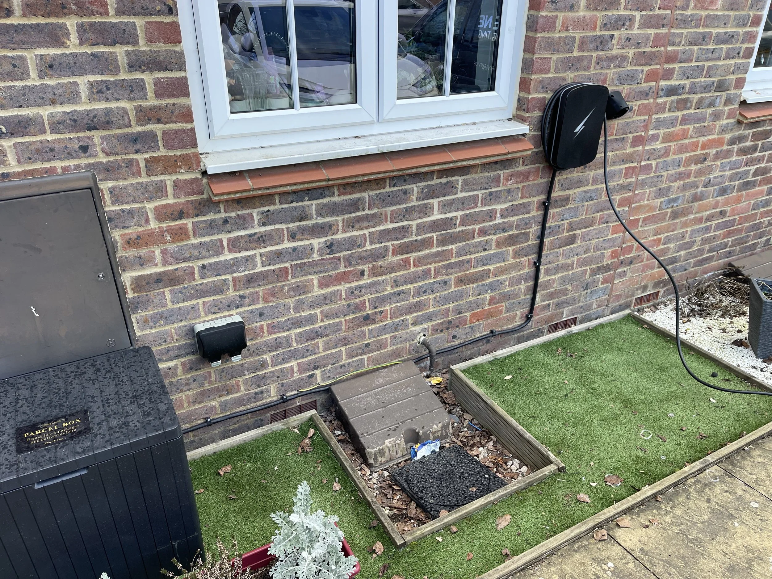 A garden area outside a brick house with a window, an electric box, and a black cable reel with cable hanging down. There are two sections of artificial grass, a small planter, and a paved pathway.