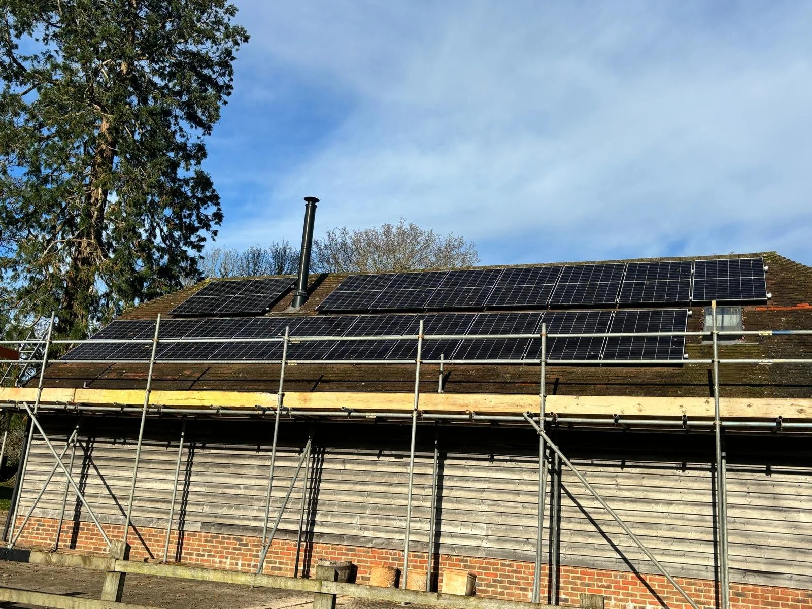 House roof with multiple solar panels installed, scaffolding around the house, under a blue sky with some clouds and trees in the background.
