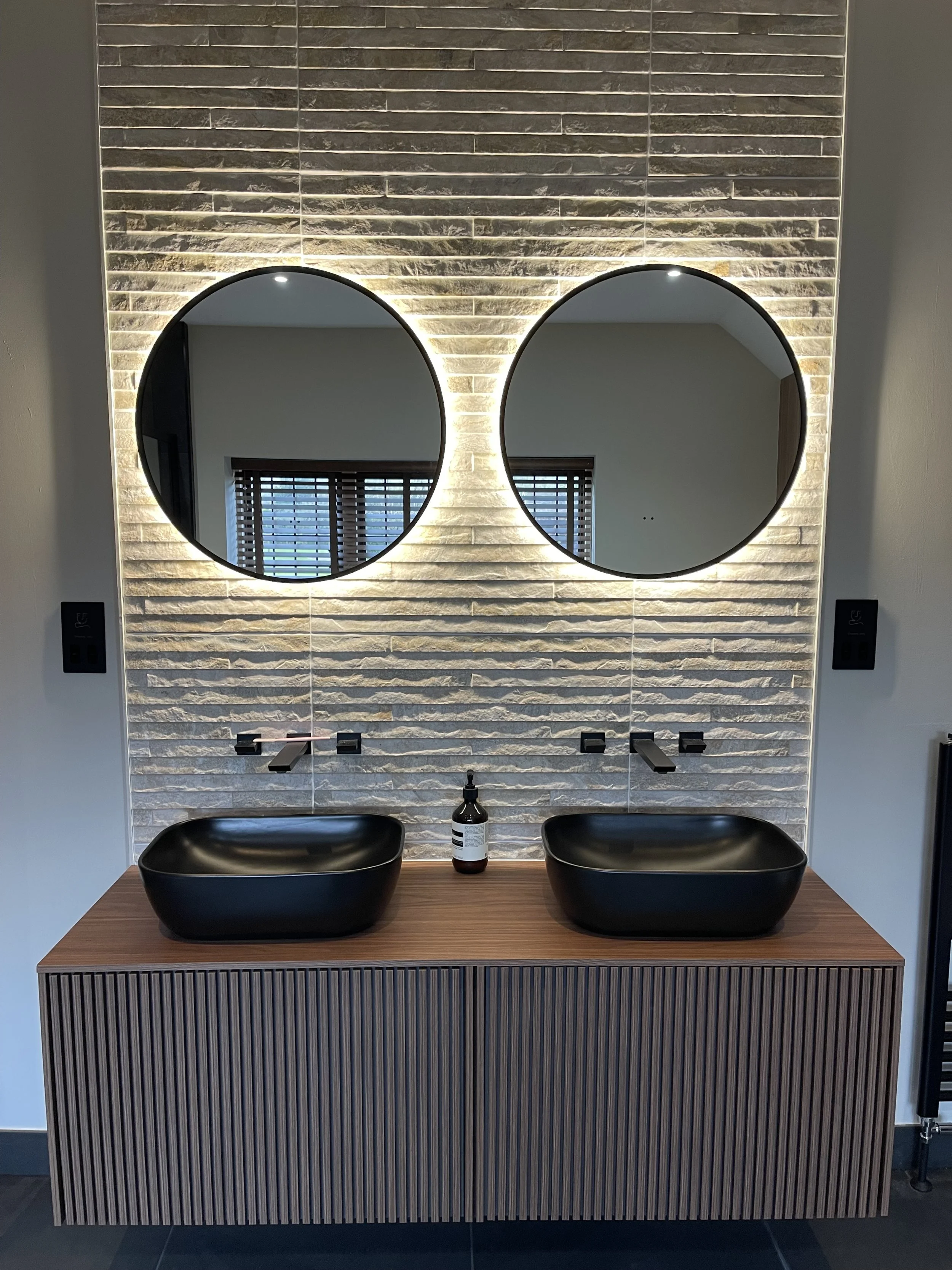 Modern bathroom vanity with two black vessel sinks, large round backlit mirrors, a wood cabinet, and a dispenser between the sinks. Reflects window with blinds.