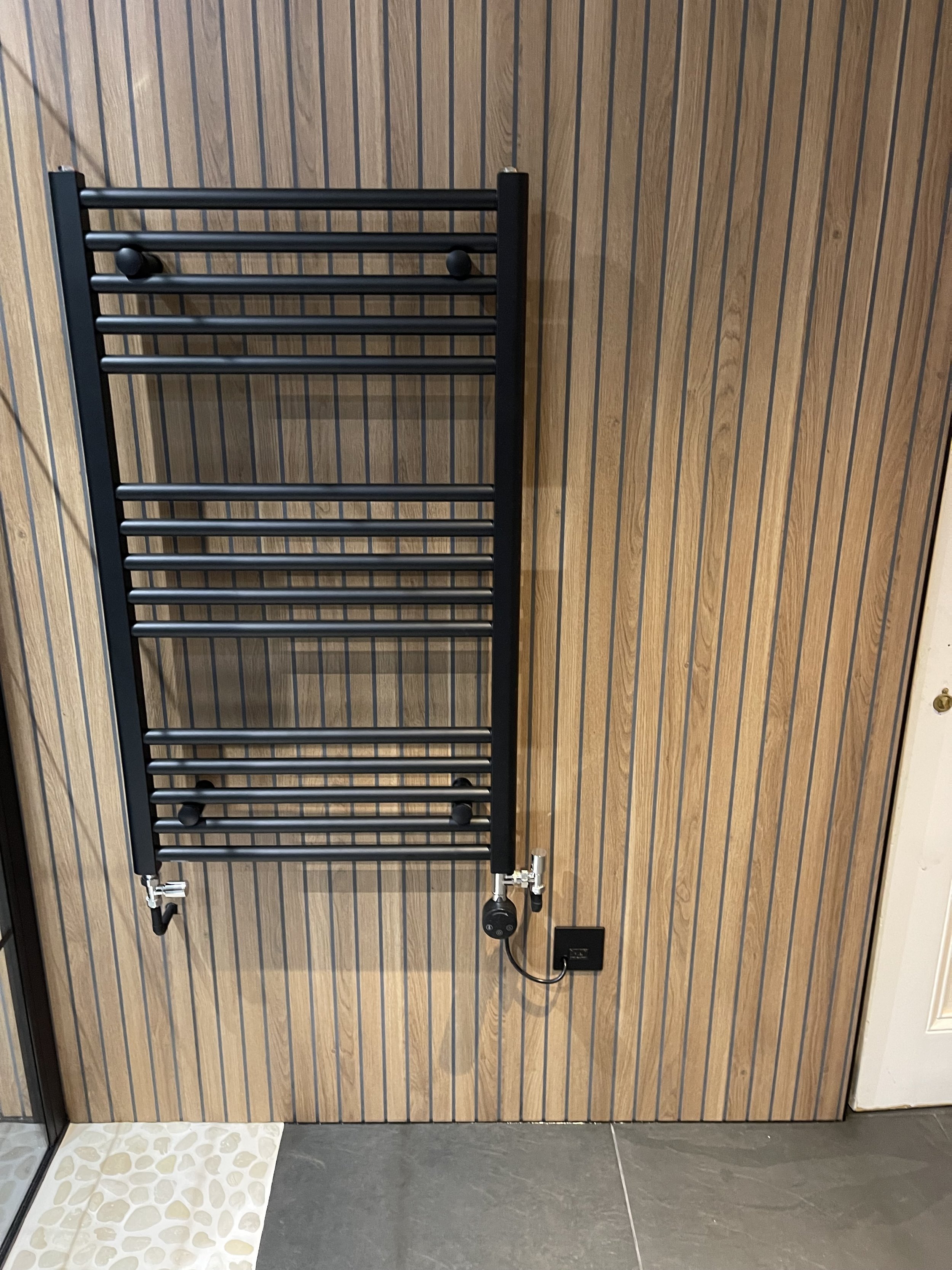 Black wall-mounted heated towel rack with multiple bars on a wood-paneled wall with an electrical plug and cord.